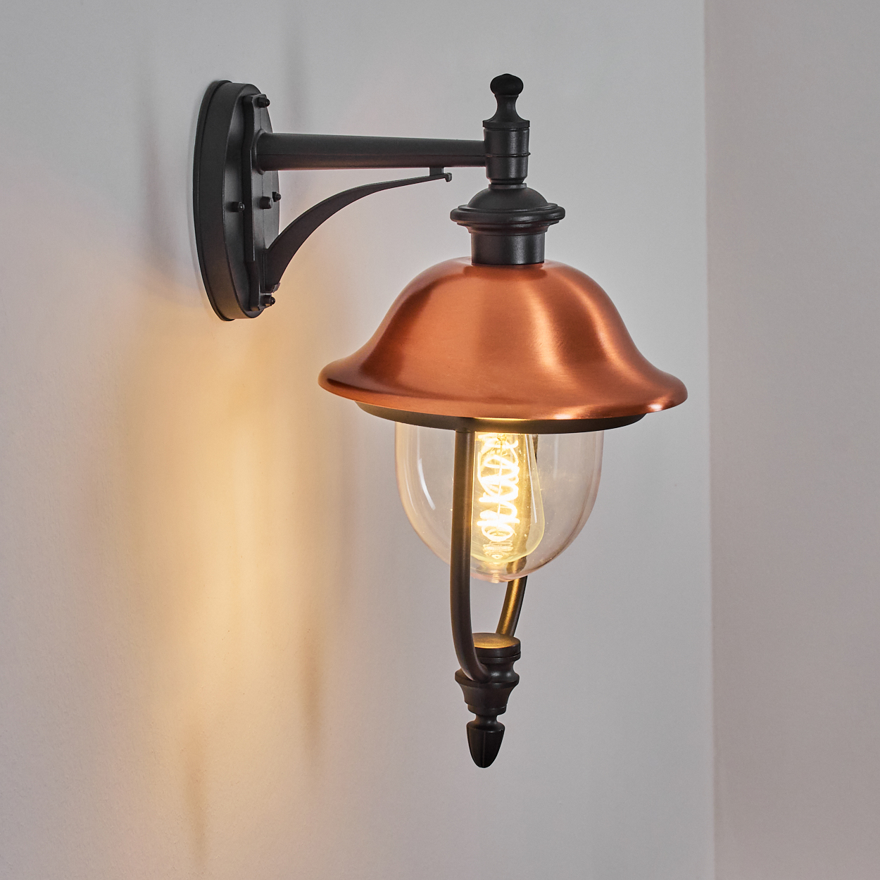 Eine an der Wand befestigte Außenlampe aus Kupfer und Schwarz. Sie hat eine Glühbirne in einem Glasgehäuse. Die Wand ist weiß.