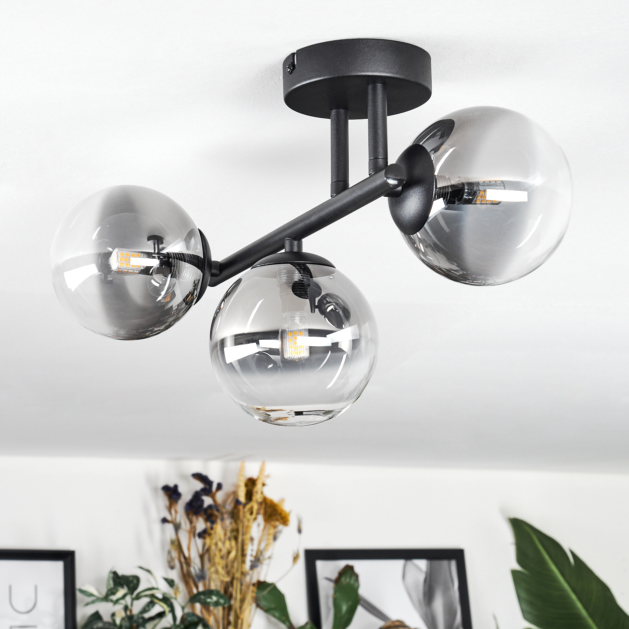 Moderne Deckenlampe mit drei kugelförmigen Glasschirmen. Schwarze Metallstruktur. Sichtbare Glühbirnen in den Schirmen. Weißer Deckenhintergrund.