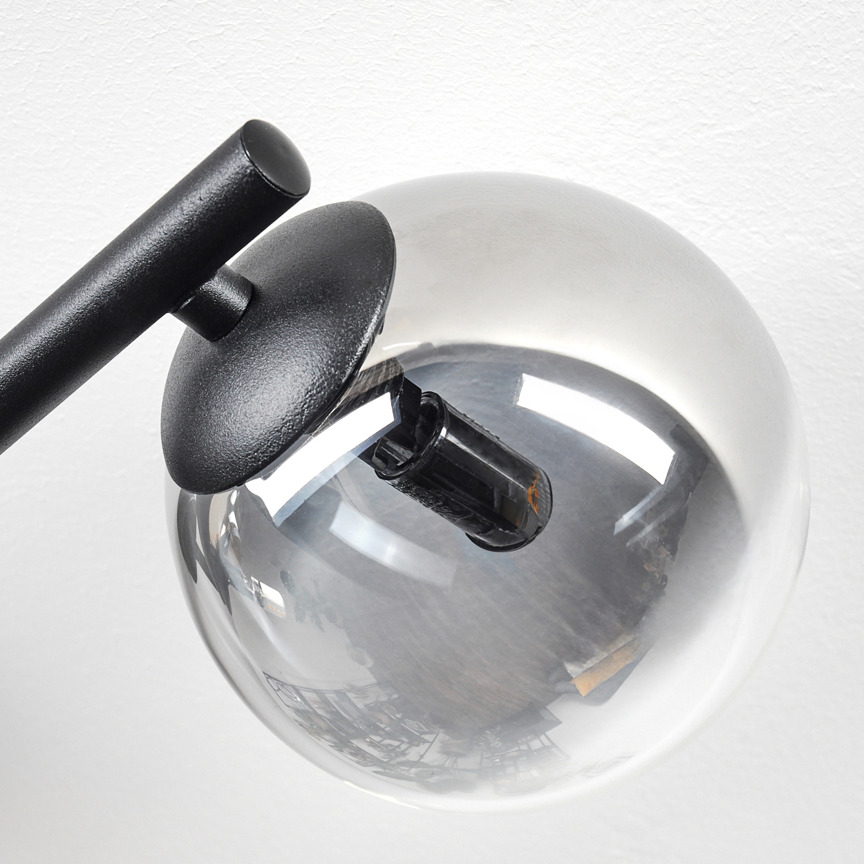 Eine moderne schwarze Lampe mit kugelförmigem Glasschirm. Sie ist an einer weißen Wand montiert. Das Glas spiegelt die Umgebung wider.