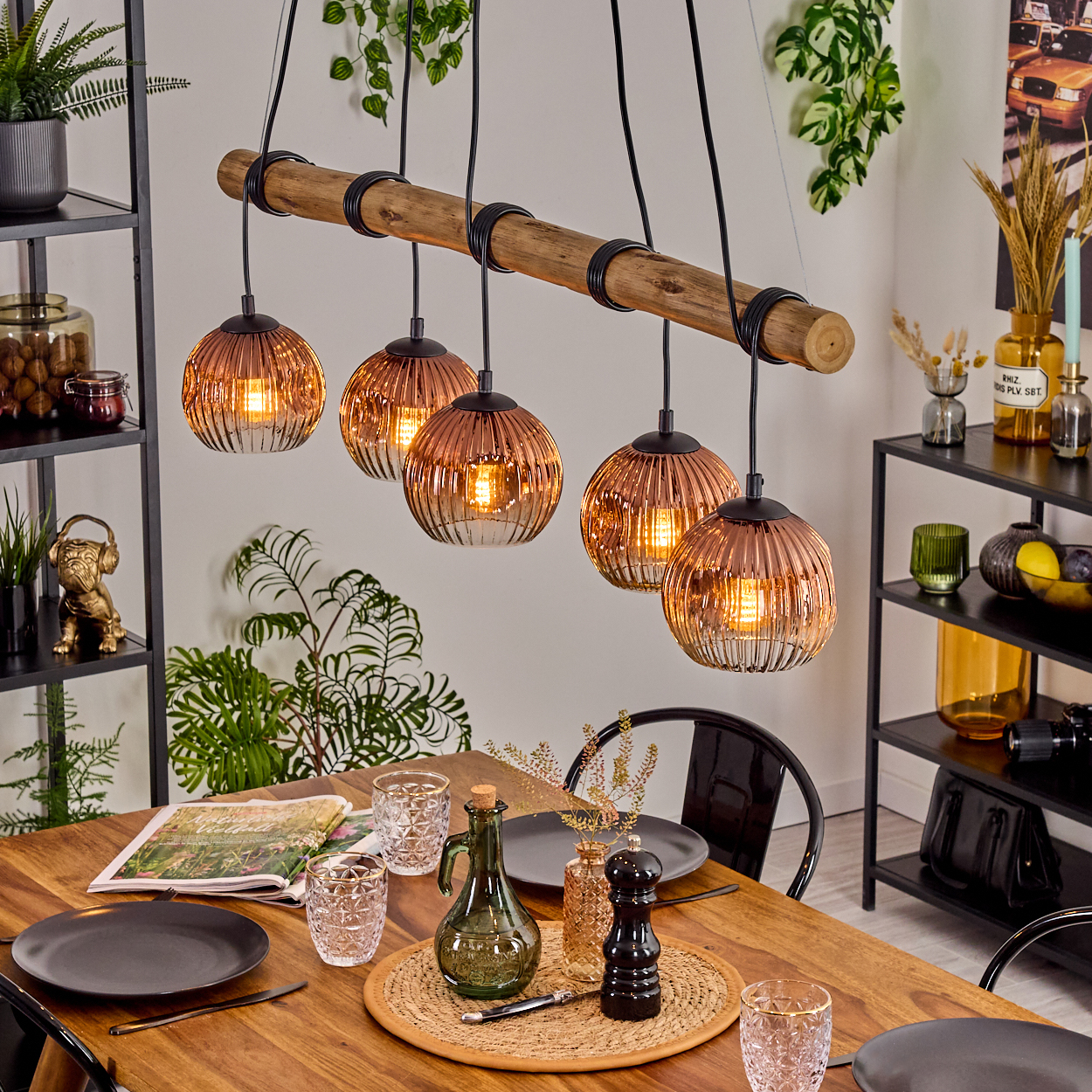 Esszimmer mit Holztisch, fünf kupferfarbenen Kugelleuchten, schwarzen Metallregalen und Dekoration. Teller und Gläser stehen auf dem Tisch.