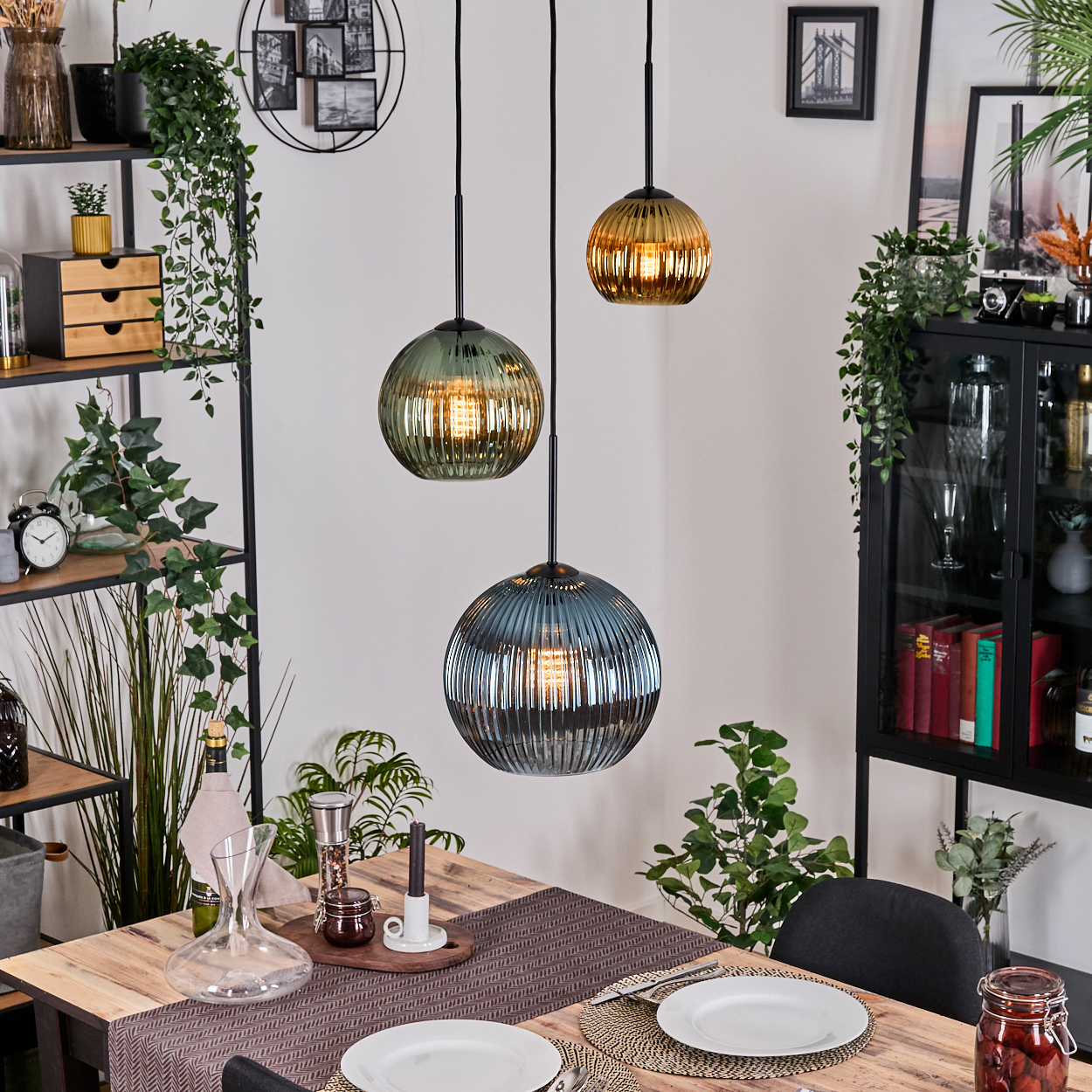 Esszimmer mit drei Hängelampen. Ein Holztisch ist mit Tellern gedeckt. Regale und ein Schrank sind im Hintergrund zu sehen.
