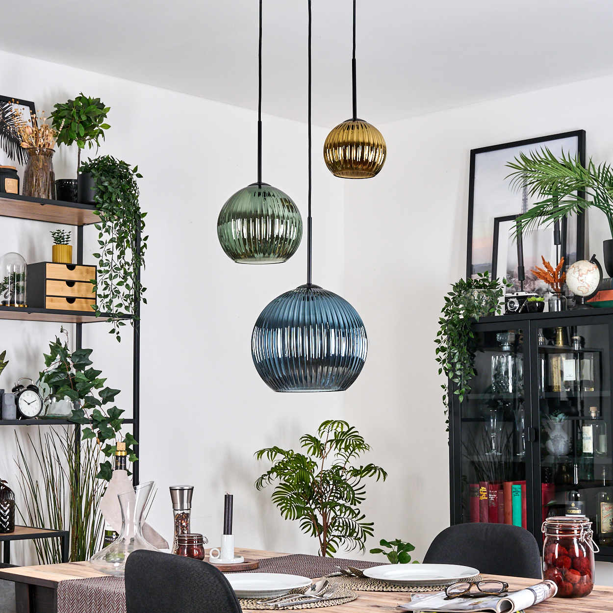 Esszimmer mit drei Hängelampen. Tisch mit Tellern. Regale mit Pflanzen und Dekor. Schwarzer Schrank mit Dekor.