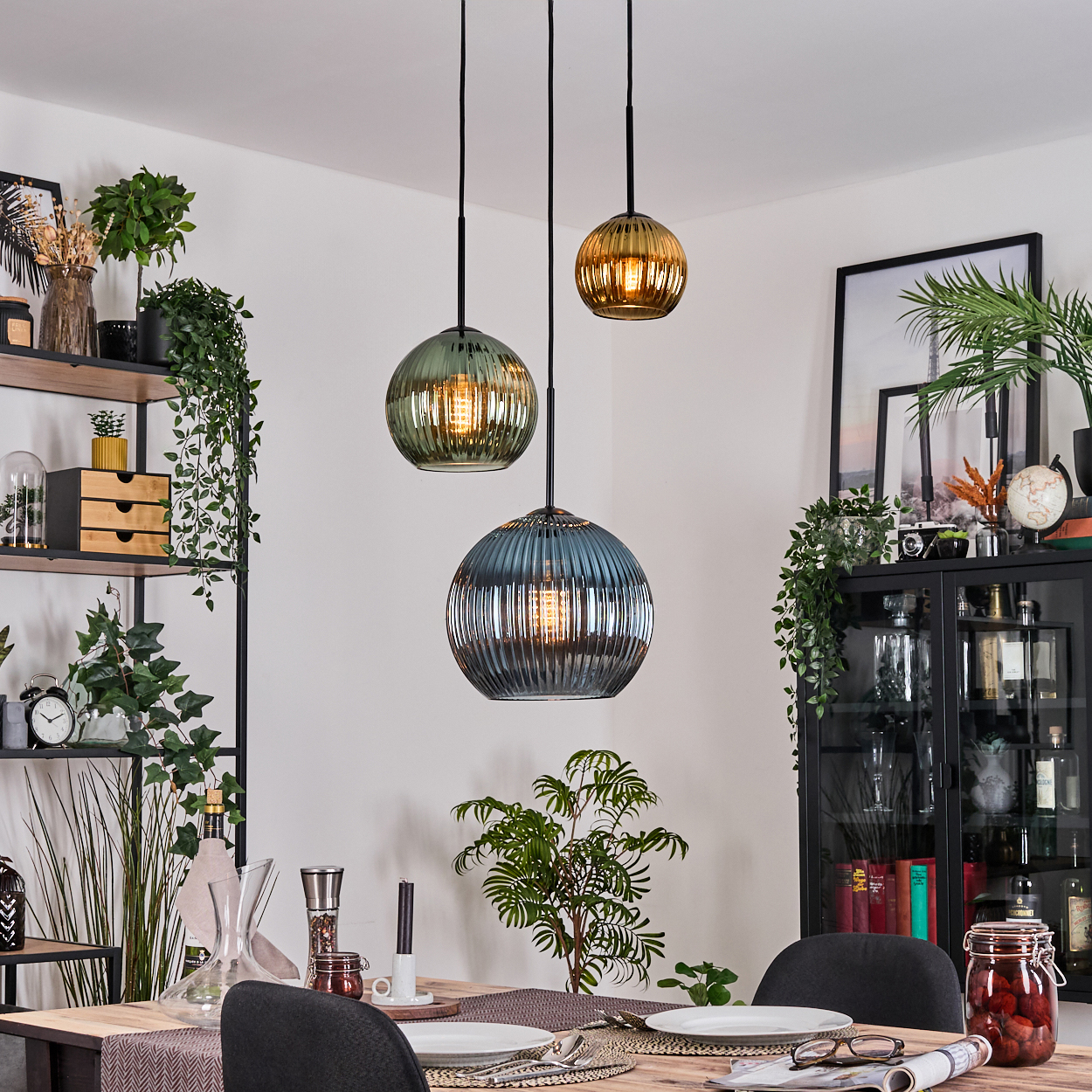 Esszimmer mit Hängelampen. Ein Tisch ist mit Tellern gedeckt. Regale und ein Schrank sind sichtbar, geschmückt mit Pflanzen und Dekor.