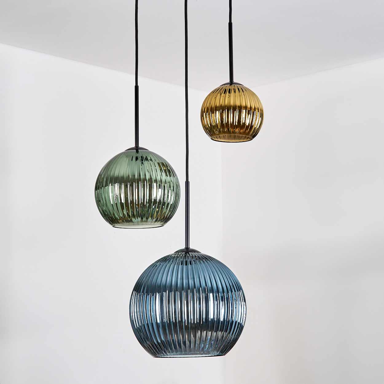Drei Pendelleuchten mit geriffelten Glasschirmen in Grün, Gold und Blau hängen vor einer weißen Wand.