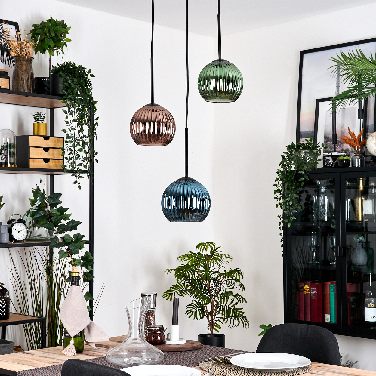 Esszimmer mit drei Hängelampen. Dekorative Pflanzen und Regale sind sichtbar. Ein Tisch ist mit Tellern und Gläsern gedeckt.