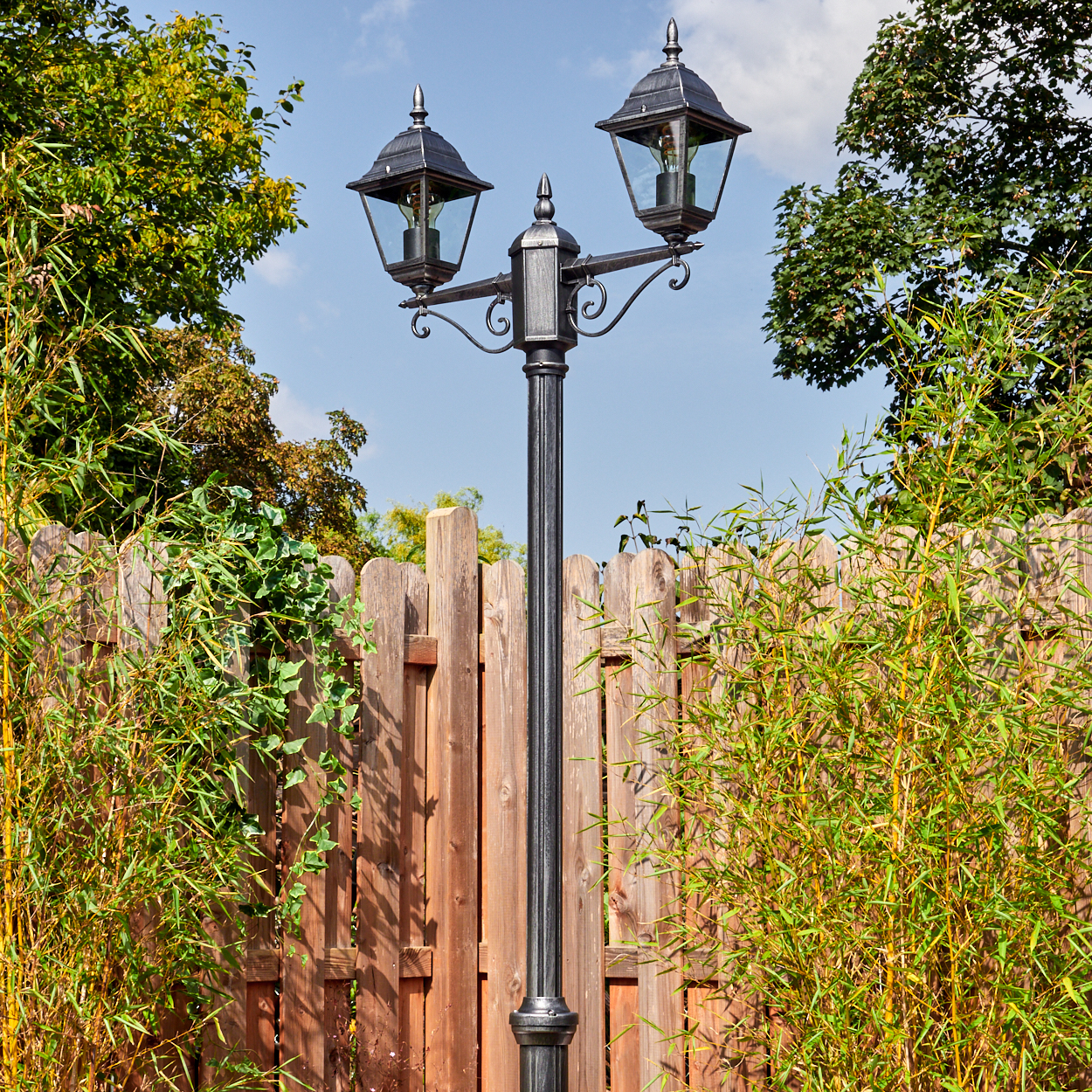 Eine schwarze Laterne mit zwei Lampen steht vor einem Holzzaun. Grüne Pflanzen und Bäume umgeben die Szene unter blauem Himmel.