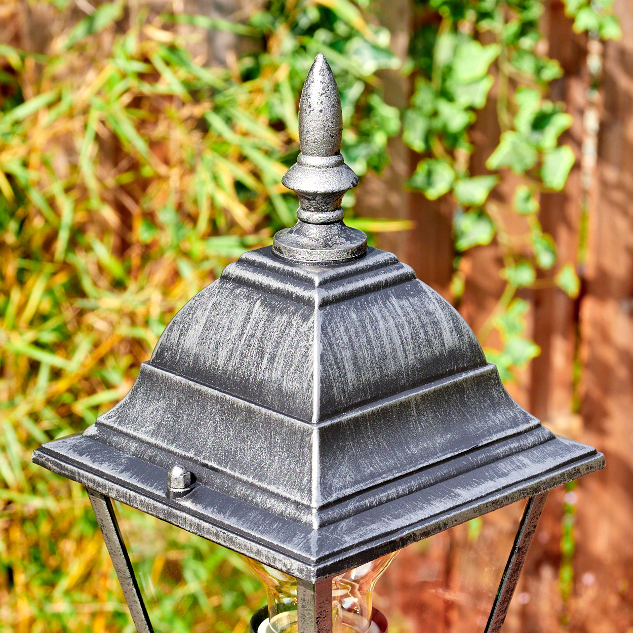 Nahaufnahme einer silbernen Außenlampe mit spitzem Aufsatz. Grünes Laub und ein Holzzaun im unscharfen Hintergrund.