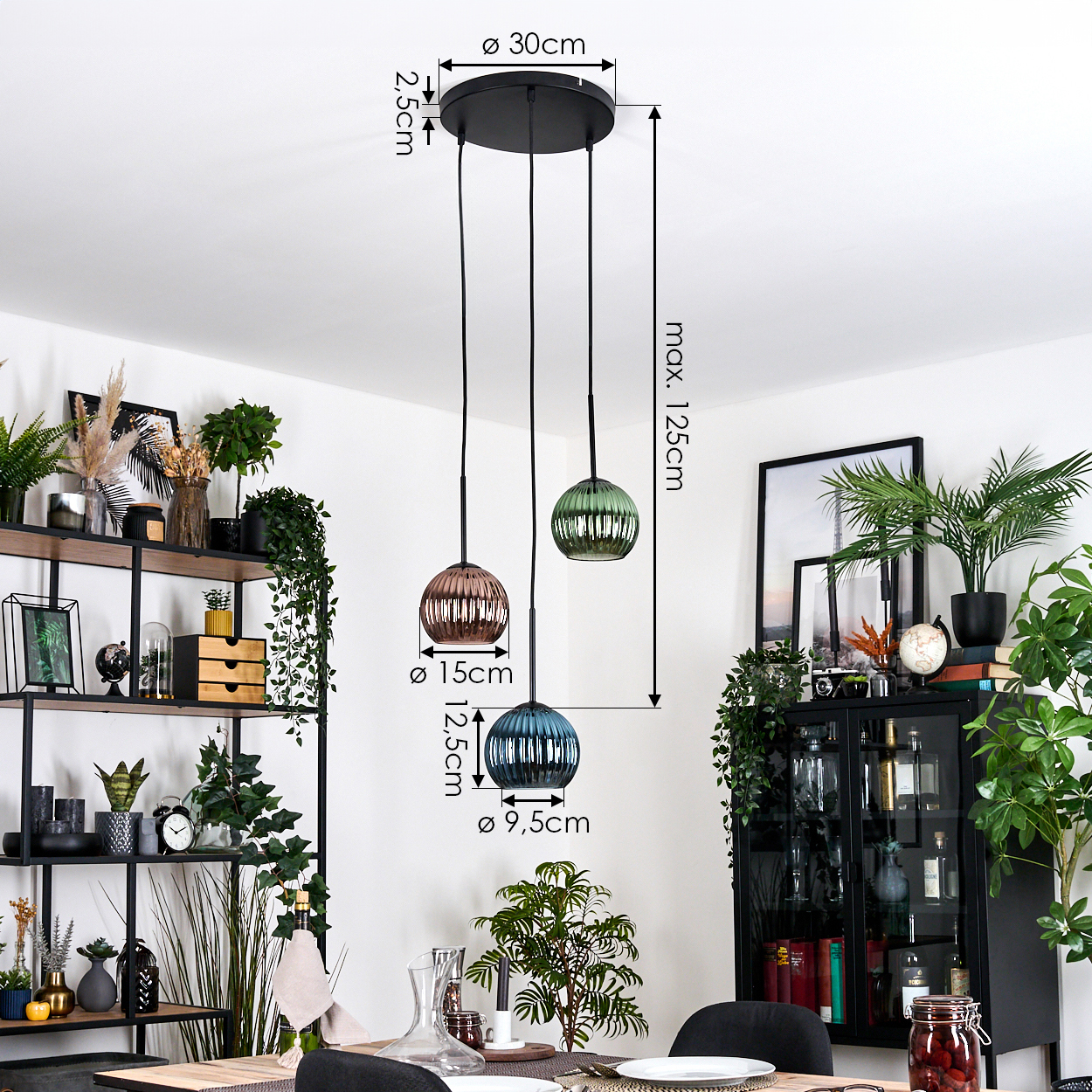 Drei Pendelleuchten mit Glasschirmen hängen von der Decke. Ein schwarzes Regal und ein Schrank sind sichtbar, gefüllt mit Pflanzen und Dekor.
