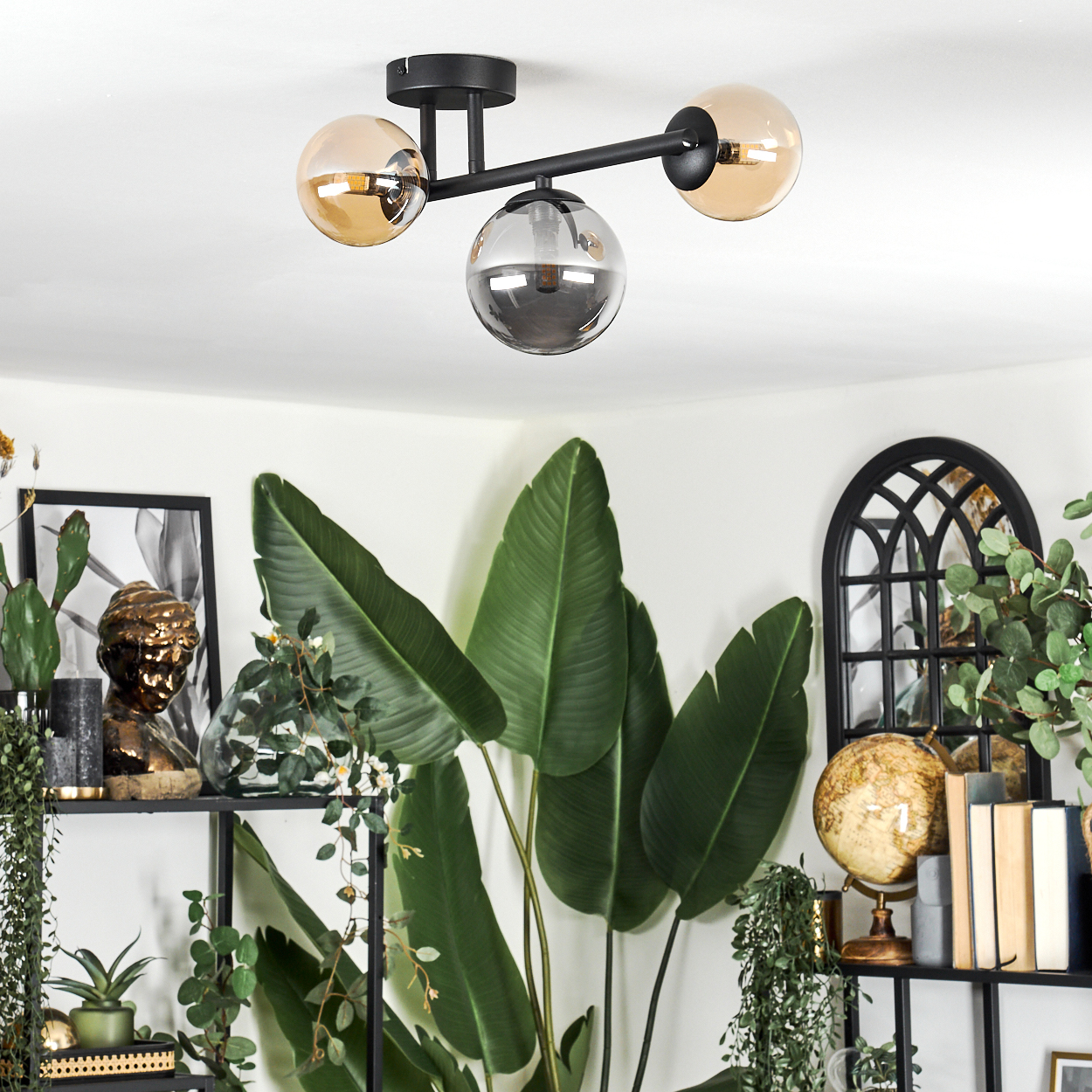 Eine moderne Deckenlampe mit drei Kugelleuchten. Darunter befinden sich Pflanzen, ein gerahmtes Bild und ein Globus auf einem Regal.