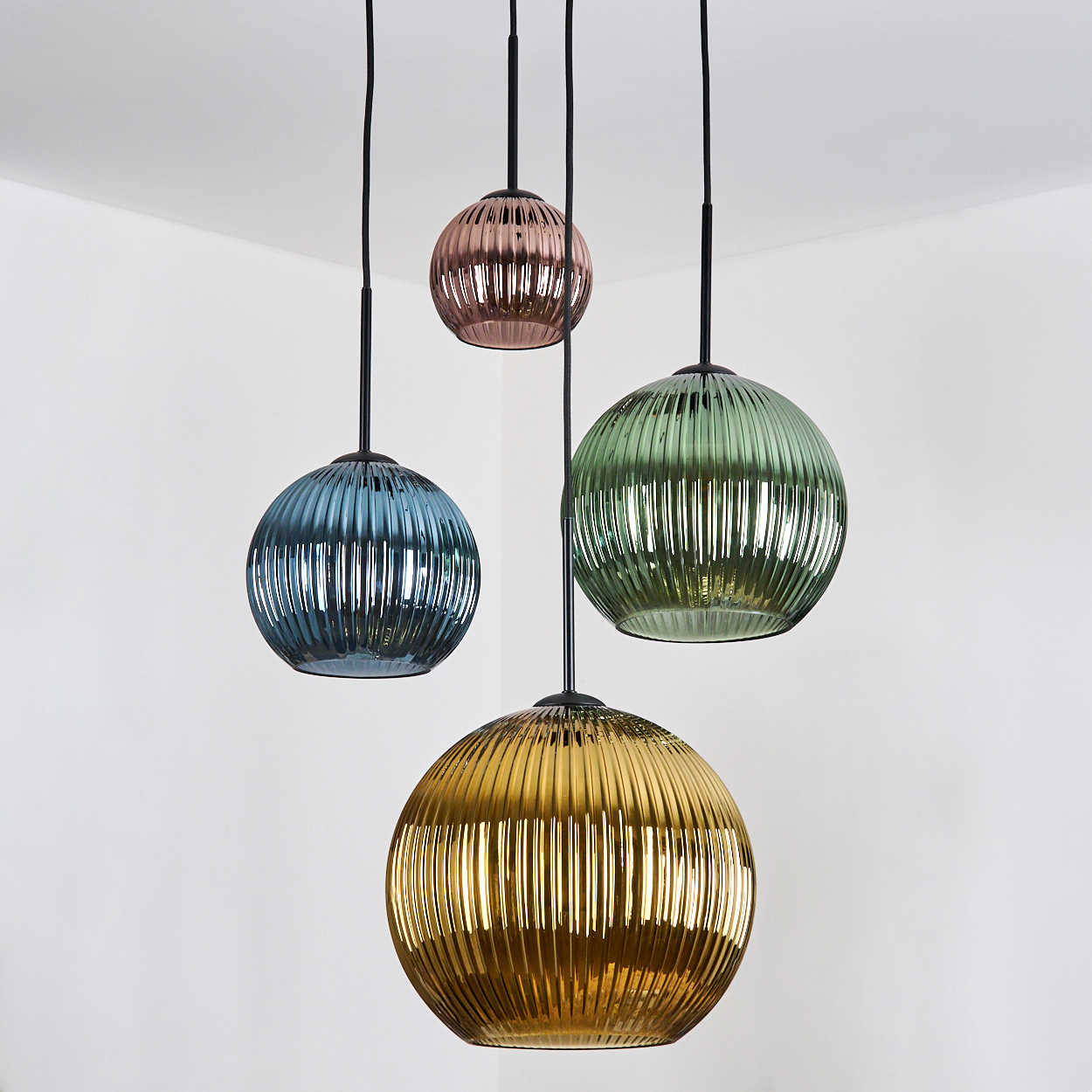 Vier bunte, gerippte Glas-Pendelleuchten hängen an schwarzen Kabeln vor einer weißen Wand. Farben sind Blau, Rosa, Grün und Gold.