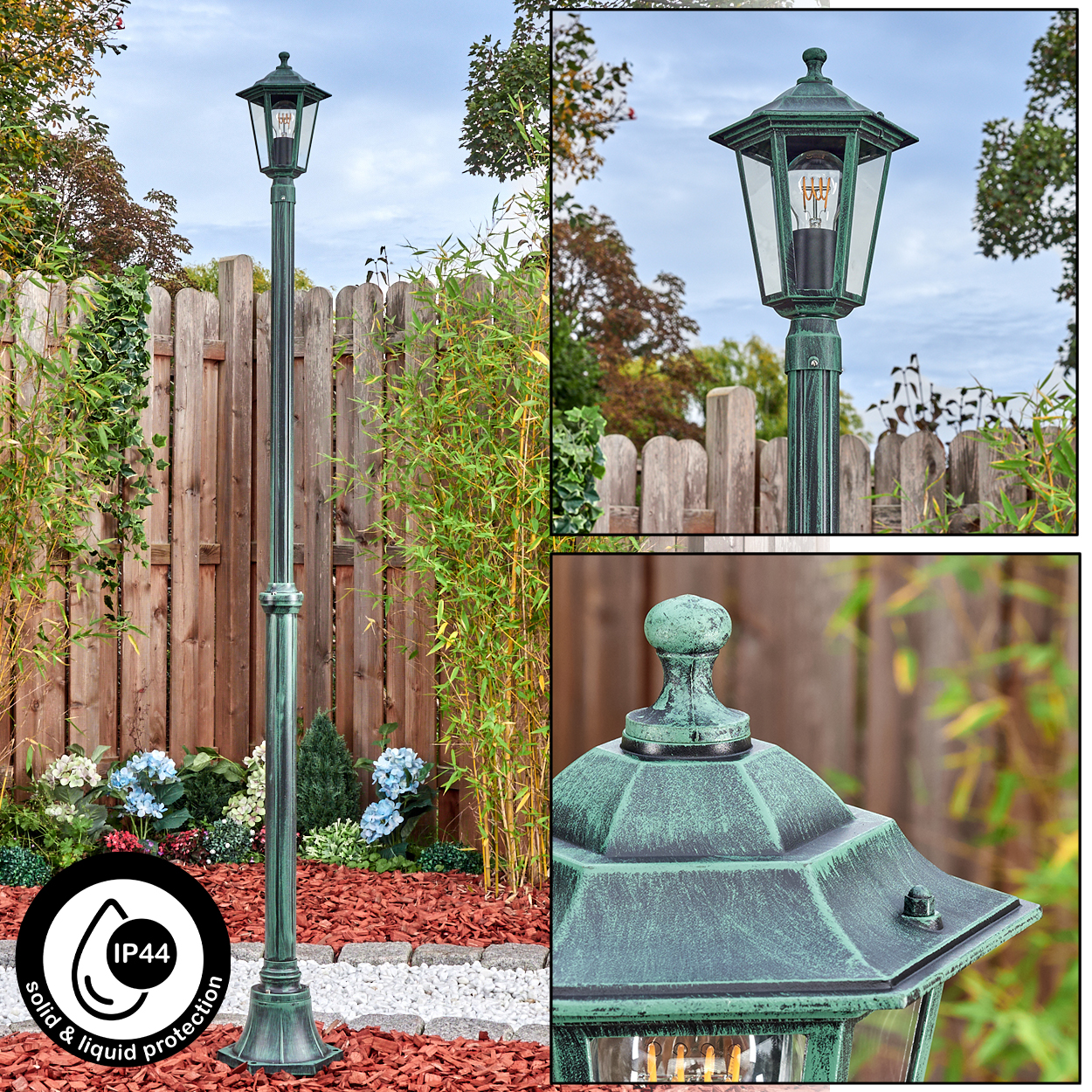 Eine grüne Außenlaterne steht in einem Garten. Im Hintergrund ist ein Holzzaun. Bambus und andere Pflanzen umgeben die Lampe.