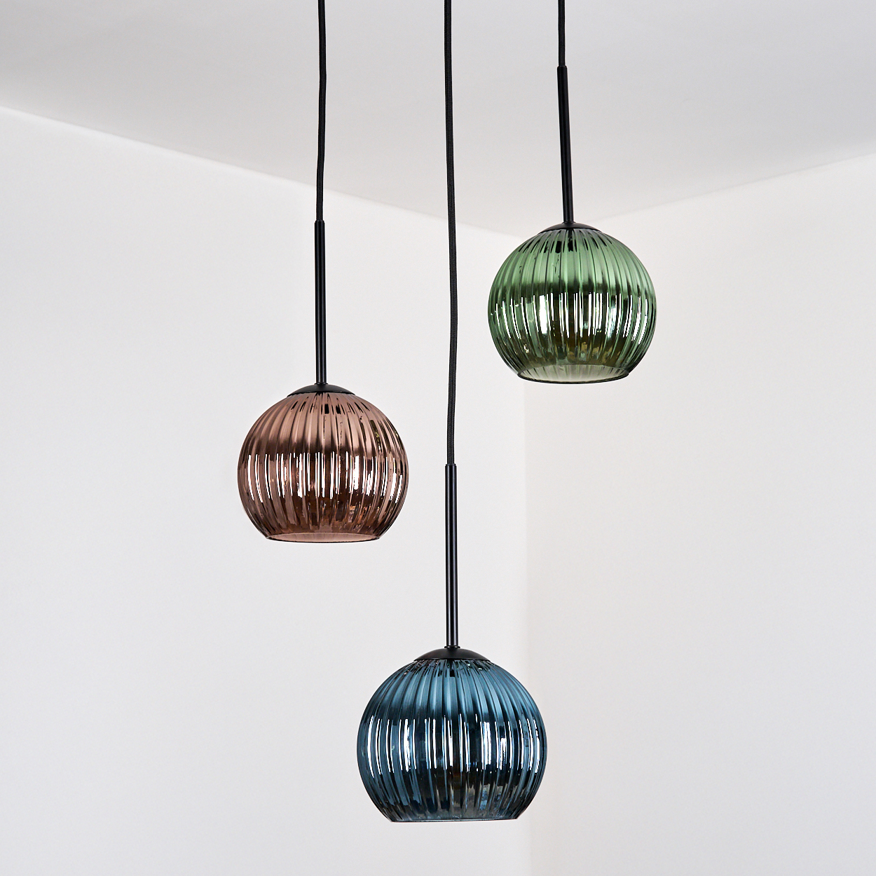 Drei Pendelleuchten mit gerippten Glasschirmen in Kupfer, Grün und Blau hängen vor einer weißen Ecke.