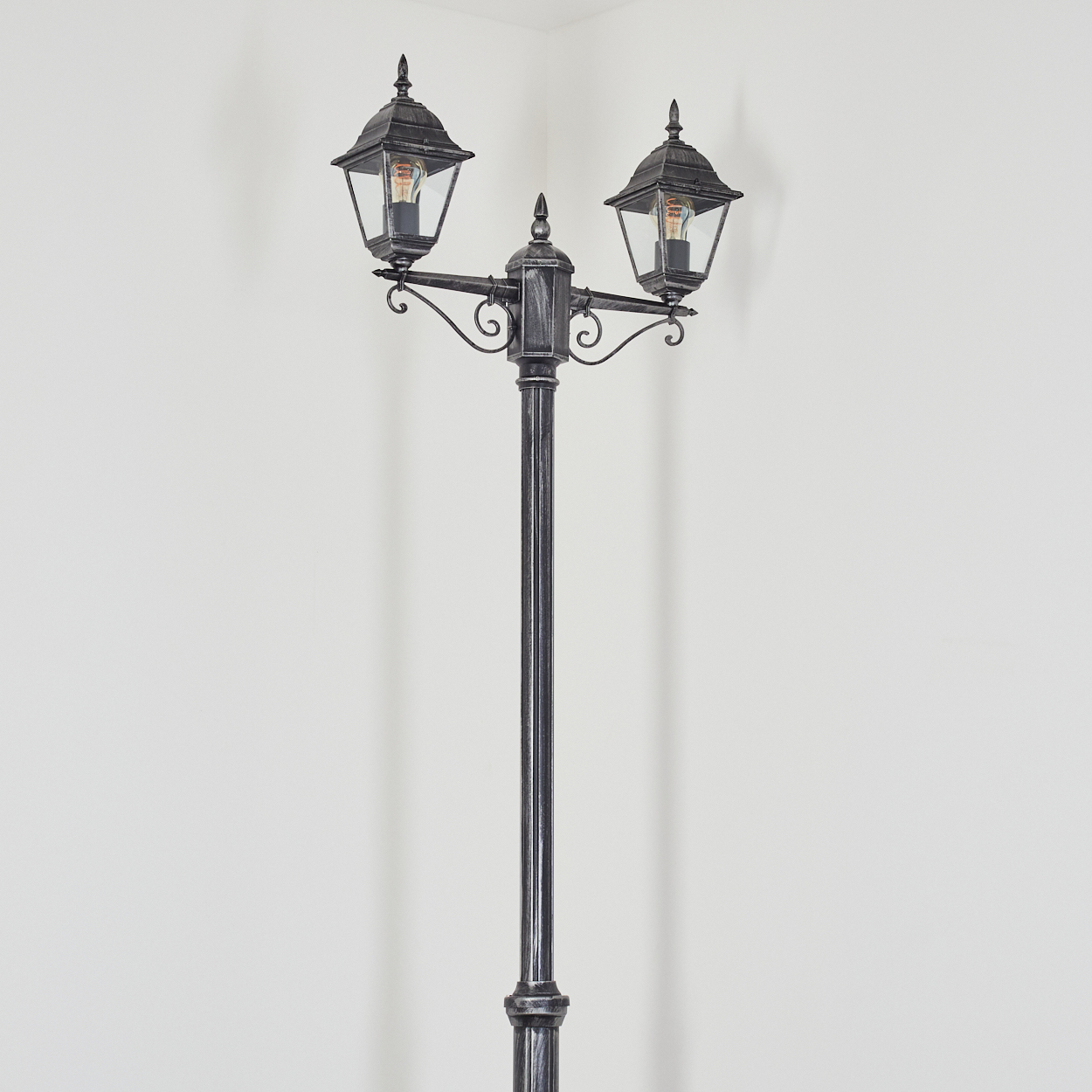 Eine dekorative schwarze Straßenlaterne mit zwei Lampen. Sie steht in einer Ecke vor einer weißen Wand. Jede Lampe hat eine sichtbare Glühbirne.