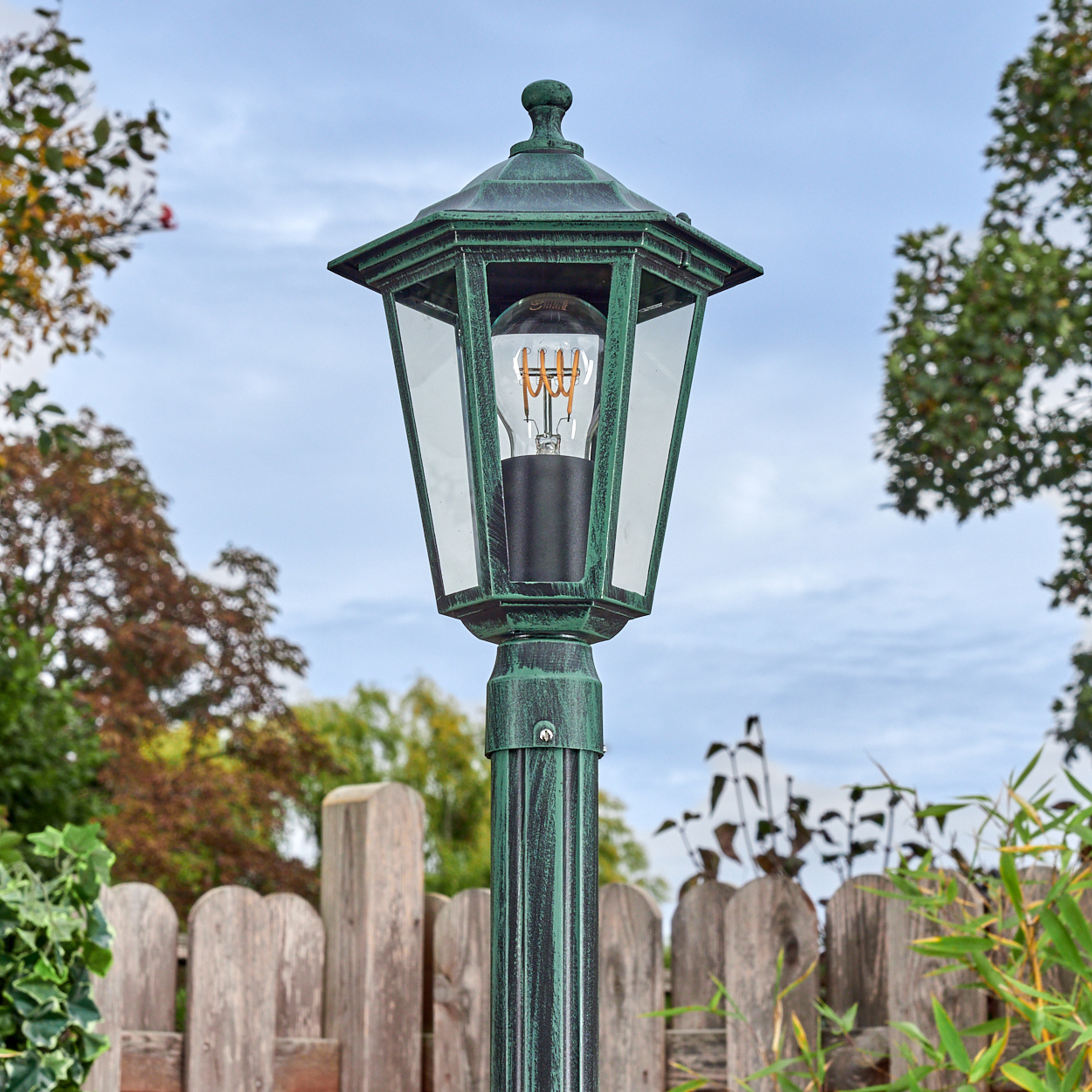 Grüne Außenlampe mit Glühbirne, vor einem Holzzaun und Bäumen, unter einem bewölkten Himmel.