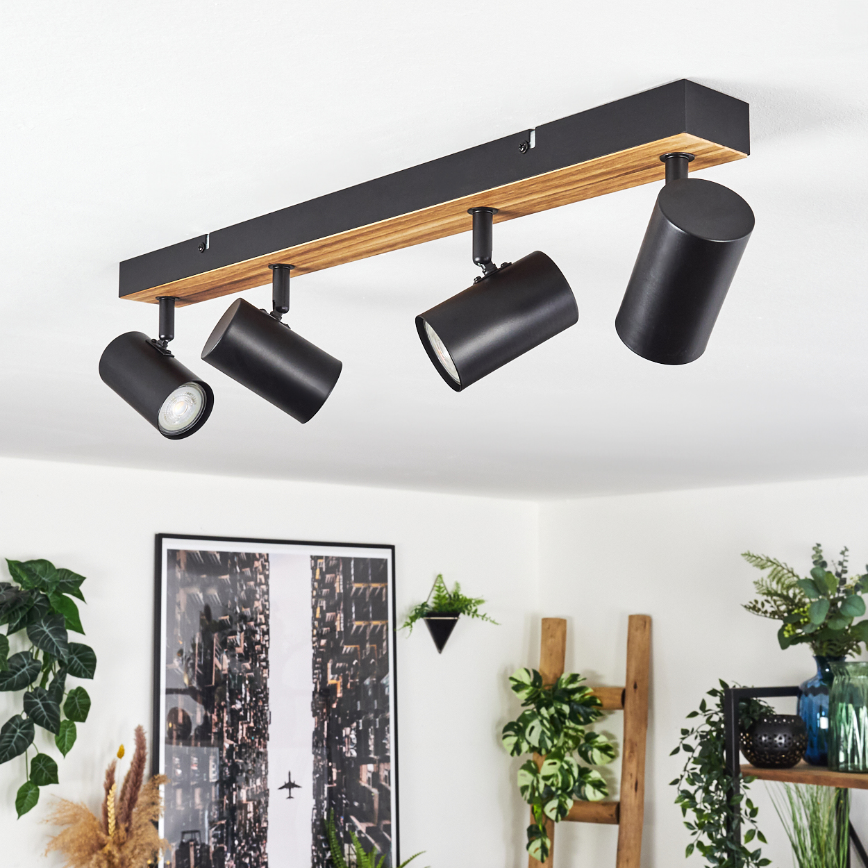 An der Decke montierte schwarze Strahler auf einem Holzbalken. Pflanzen und ein gerahmtes Bild im Hintergrund. Weiße Decke.
