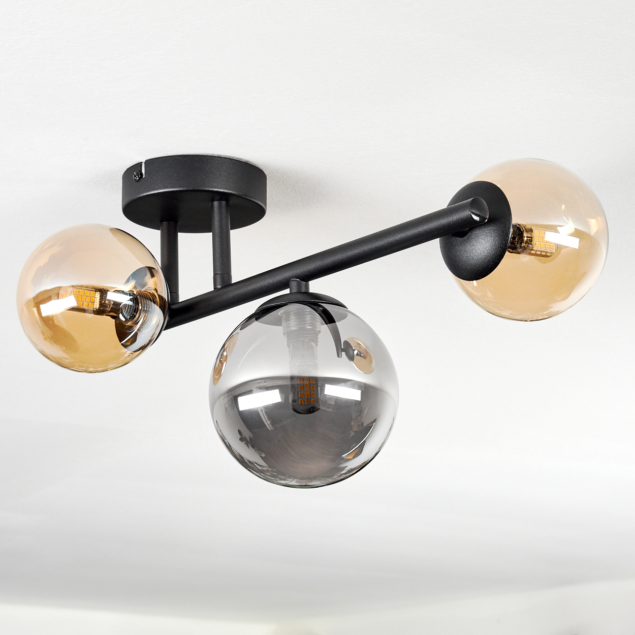 Eine moderne Deckenlampe mit drei kugelförmigen Schirmen. Die Schirme sind bernsteinfarben und grau, die Halterung schwarz.