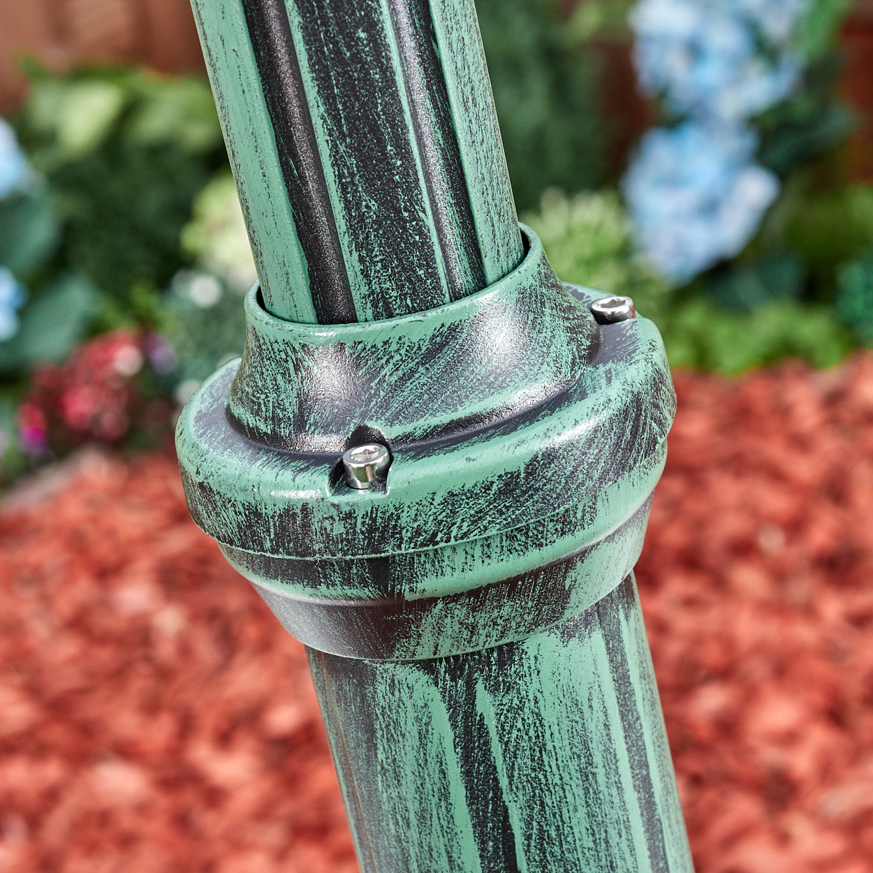 Nahaufnahme eines grünen, strukturierten Metallstabs mit Bolzen, vor verschwommenem Gartenhintergrund mit roter Mulche.