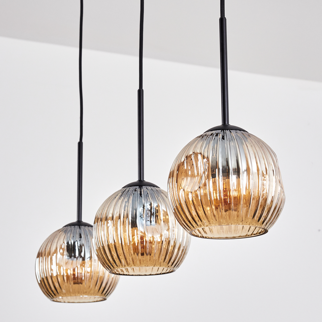 Drei Pendelleuchten mit strukturierten Glasschirmen. Schwarze Stangen und Drähte verlaufen von der Decke. Schirme sind gold und silber.