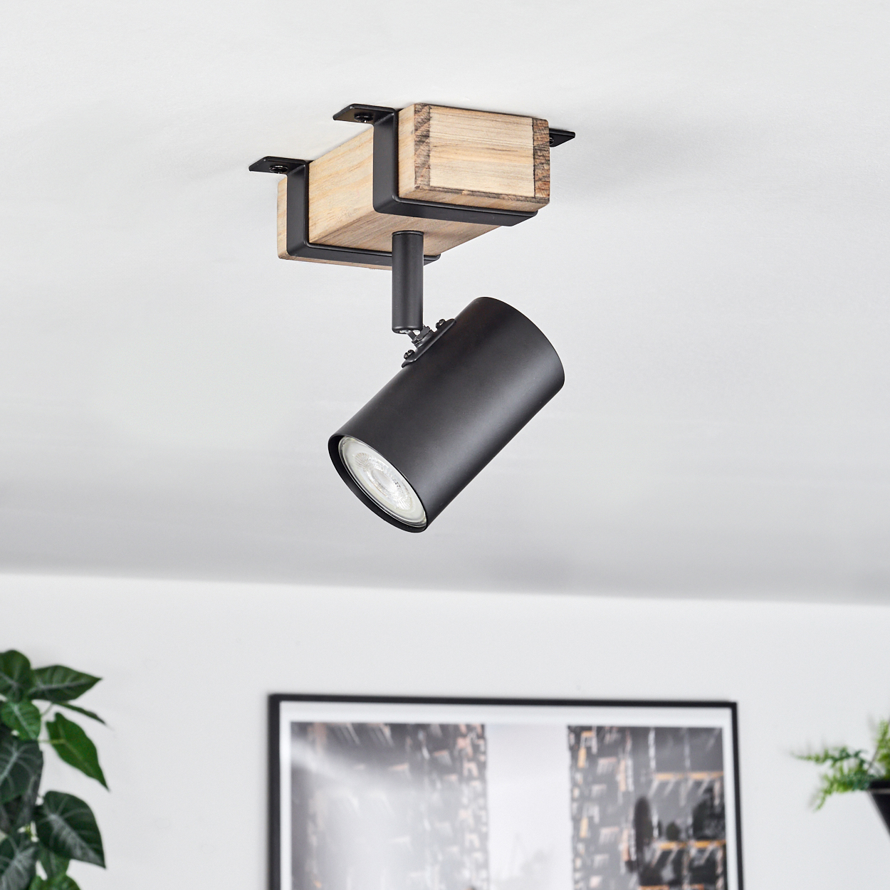 Ein Deckenstrahler mit schwarzer Lampe, Holzbalken und schwarzen Metallwinkeln. Ein gerahmtes Bild und Pflanzen im Hintergrund.