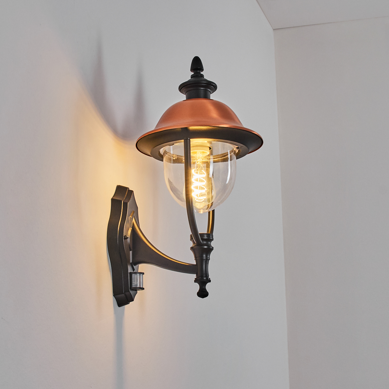 Eine Außenwandleuchte. Sie hat einen kupferfarbenen Schirm und eine durchsichtige Glühbirne. Die Lampe ist an einer weißen Wand befestigt.