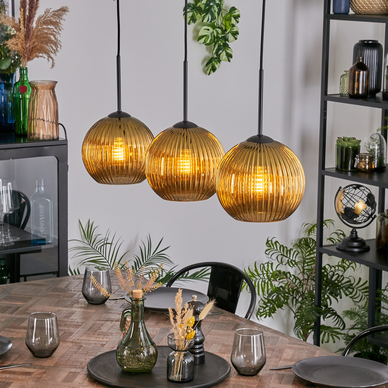 Drei goldene Pendelleuchten über einem Holztisch, der zum Abendessen gedeckt ist. Dekorative Vasen und Pflanzen im Hintergrund.