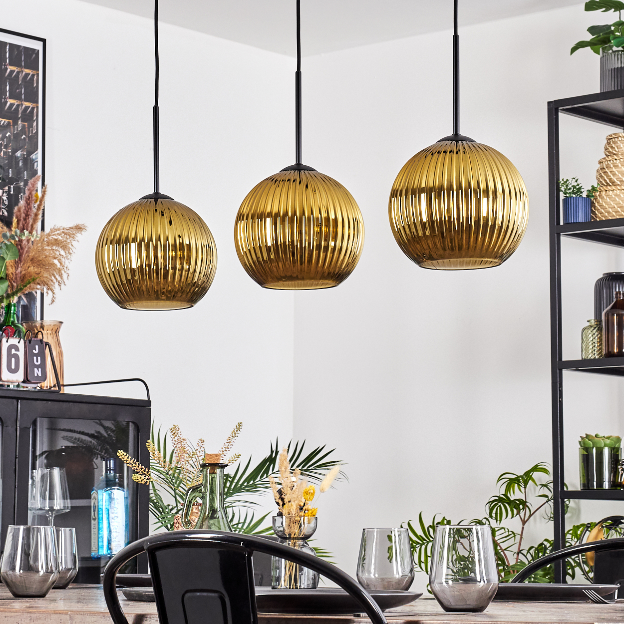 Drei goldene Pendelleuchten über einem Esstisch mit Gläsern und Pflanzen. Ein schwarzes Metallregal ist im Hintergrund.