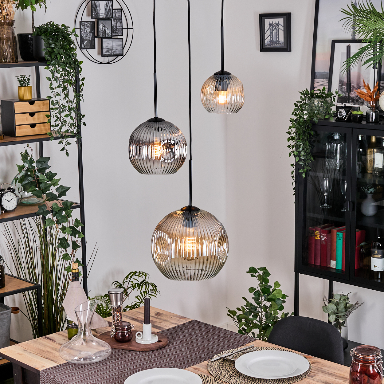 Esszimmer mit drei Hängelampen. Holztisch, Teller und Dekorationen. Regale mit Pflanzen und Dekor sind im Hintergrund sichtbar.