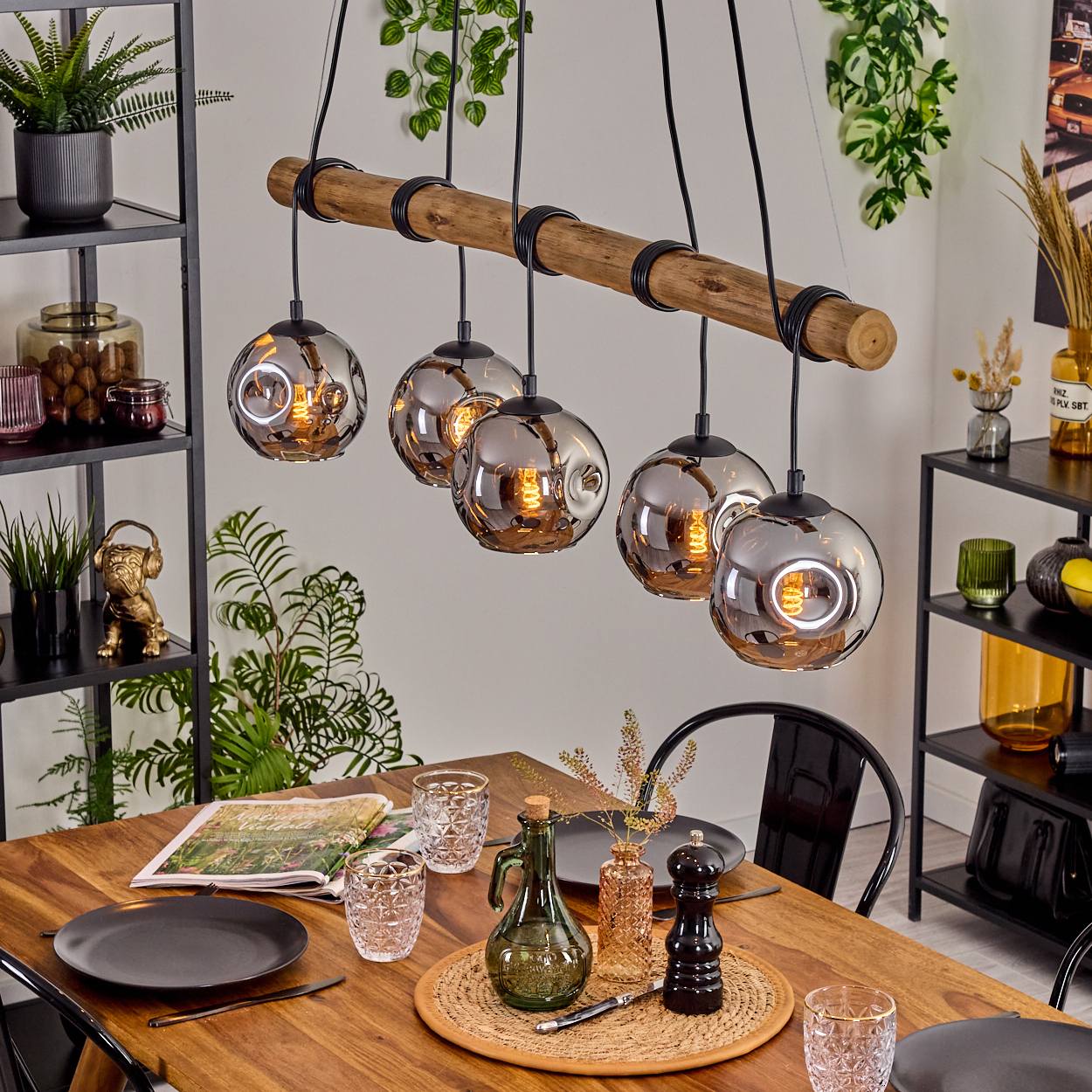 Esszimmer mit Holztisch, Glaslampen und Dekorationsartikeln. Regale mit Pflanzen und Dekor im Hintergrund.