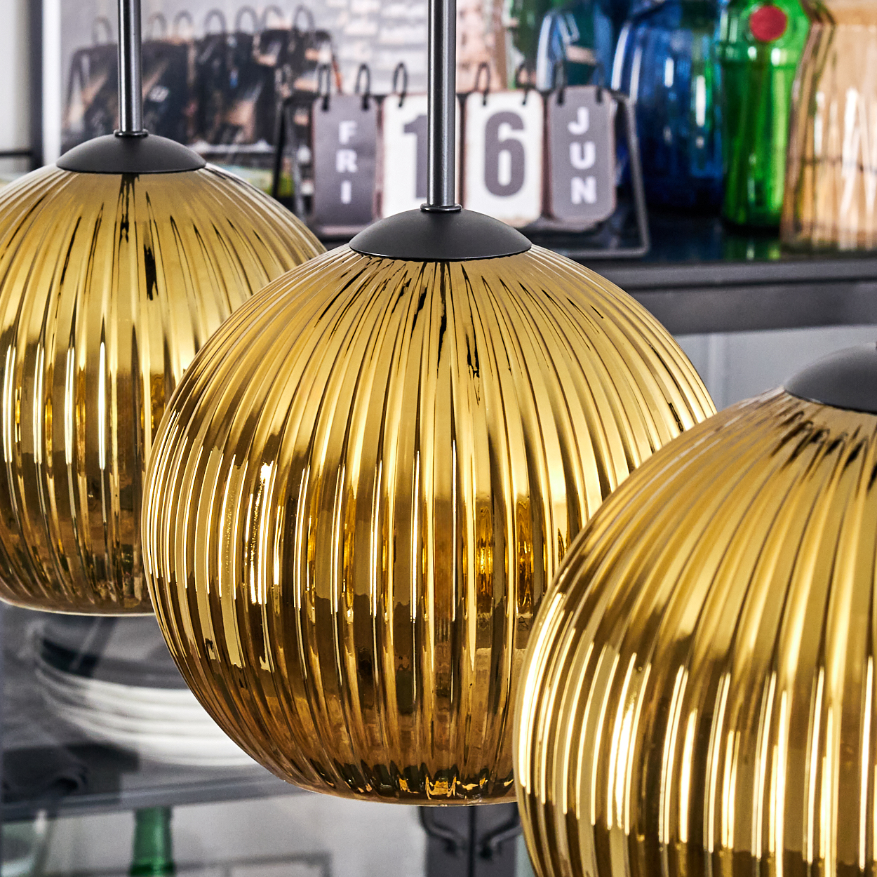 Drei goldene, gerippte Pendelleuchten hängen in einer Reihe. Ein Kalender zeigt '16 JUN'. Flaschen und Teller sind im Hintergrund.
