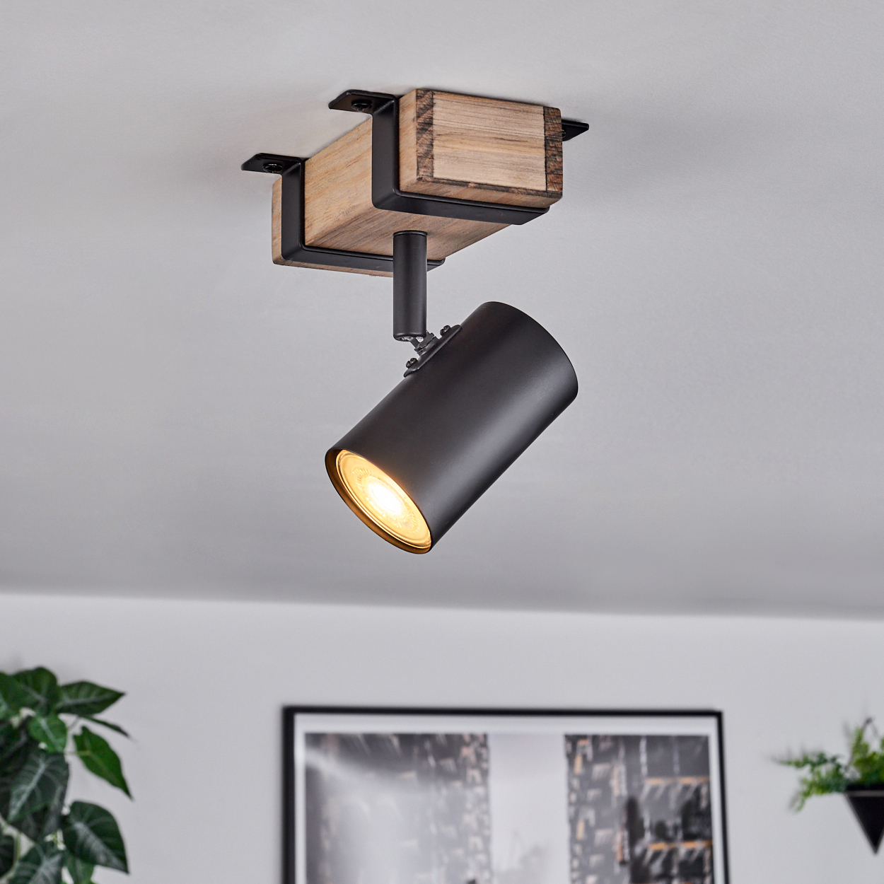 Eine schwarze Strahlerlampe ist an einem Holzbalken an der Decke befestigt. Das Licht ist an. Links ist eine grüne Pflanze zu sehen.
