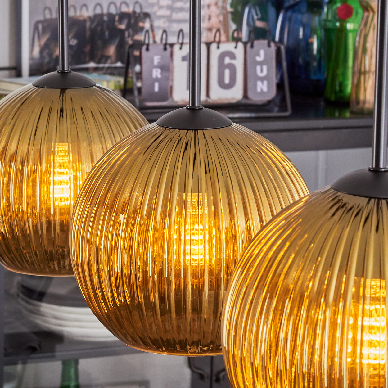 Drei goldene, gerippte Glas-Pendelleuchten mit leuchtenden Glühbirnen. Kalender im Hintergrund zeigt '16 JUN'.