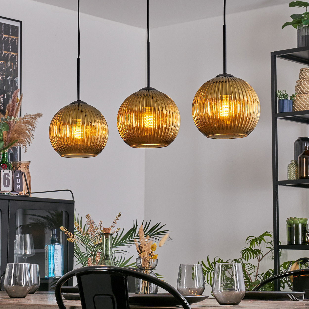 Drei goldene Pendelleuchten hängen über einem Esstisch, der mit Gläsern und Dekoration gedeckt ist. Ein schwarzes Regal und ein Schrank sind im Hintergrund.