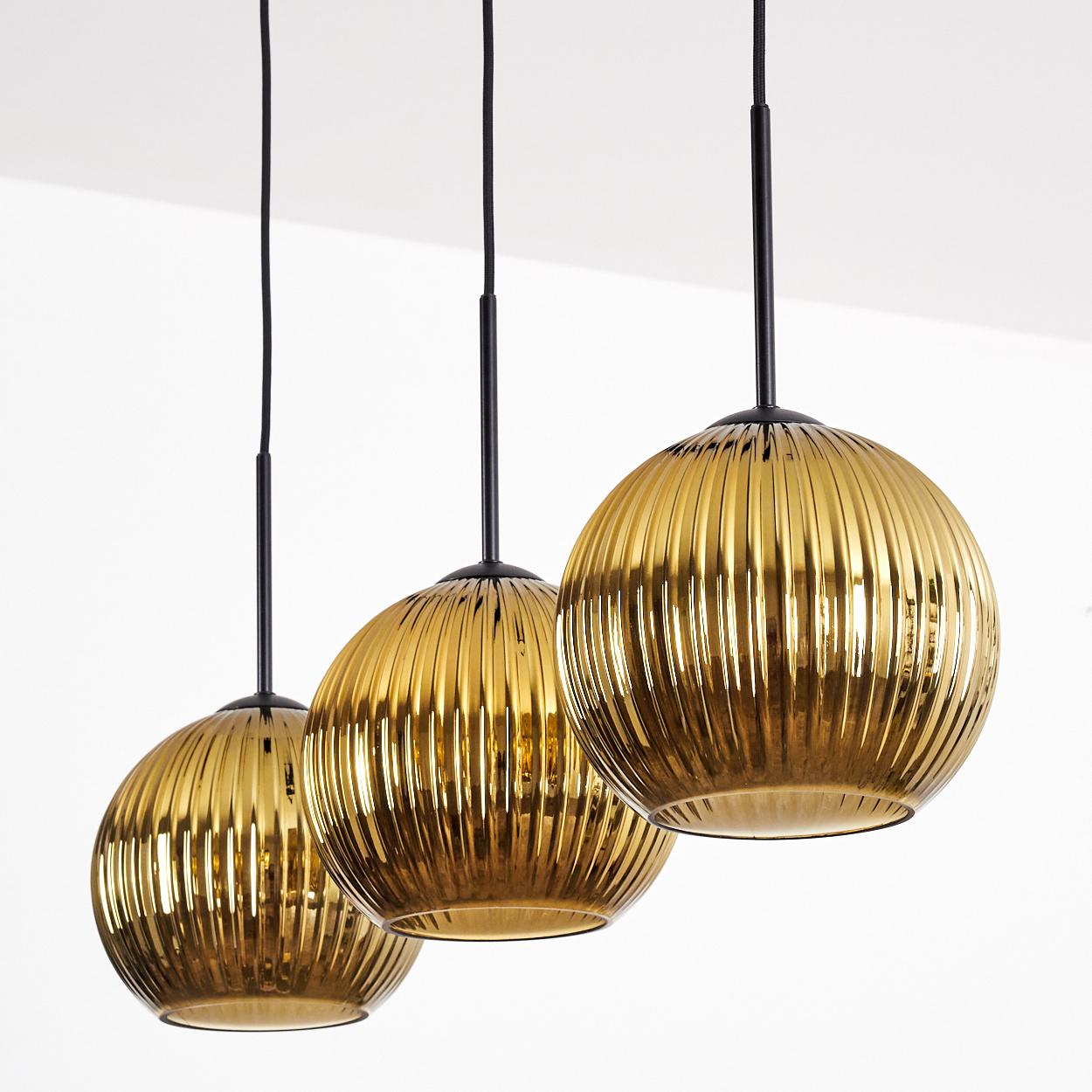 Drei goldfarbene, gerippte Glas-Pendelleuchten hängen an schwarzen Schnüren von einer weißen Decke. Die Glühbirnen haben eine kugelförmige Form.