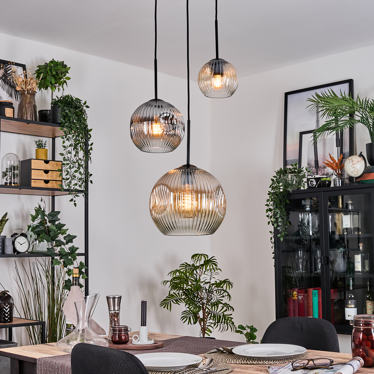 Esszimmer mit drei Hängelampen. Tisch mit Tellern, Pflanzen und Dekor. Regale und Schrank mit Pflanzen und Gegenständen.