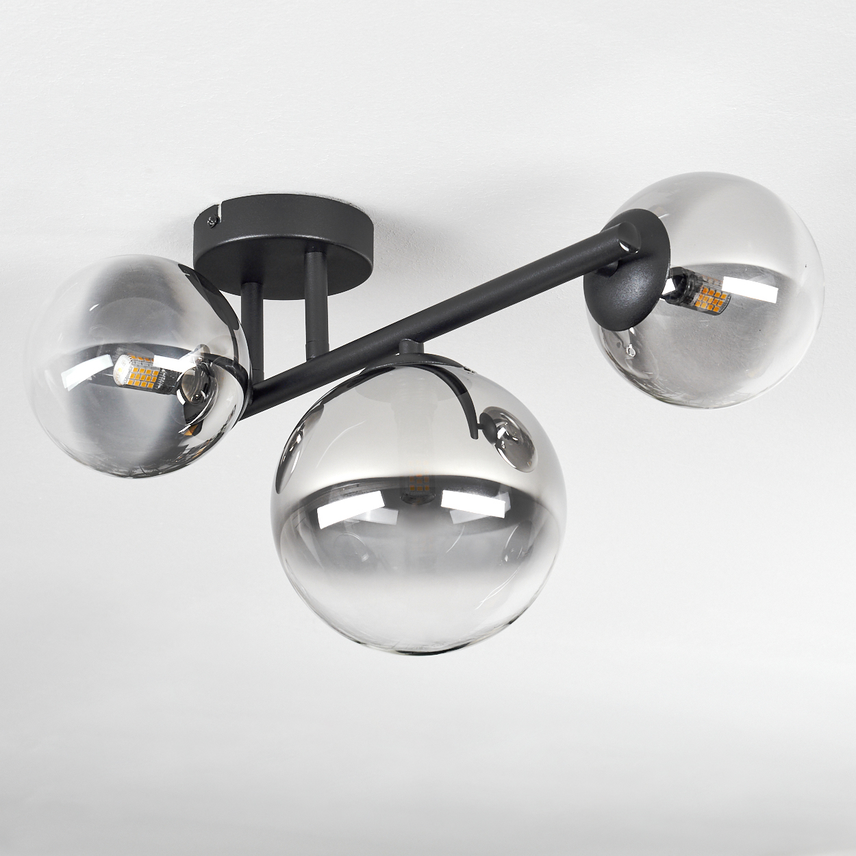 Eine Deckenleuchte mit drei kugelförmigen Glasschirmen. Die Schirme sind grau und reflektieren das Licht. Schwarze Metallarme verbinden die Schirme.