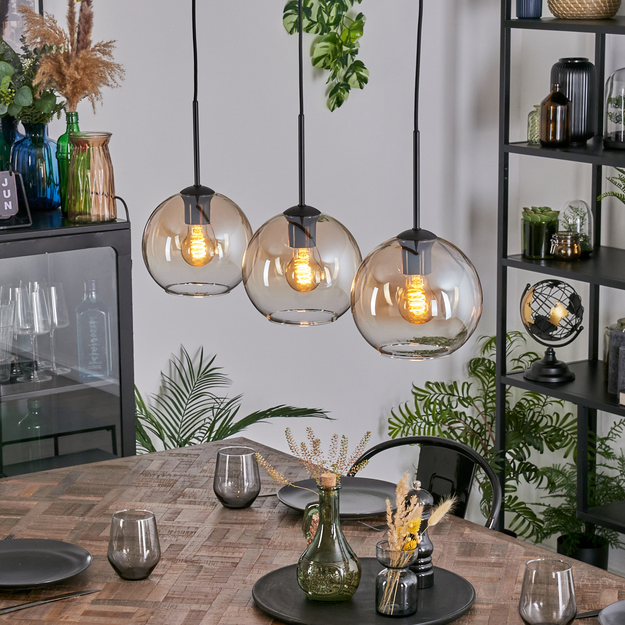 Drei Pendelleuchten über einem Holztisch mit Gläsern und Tellern. Hintergrund: Regale mit Deko und Pflanzen.