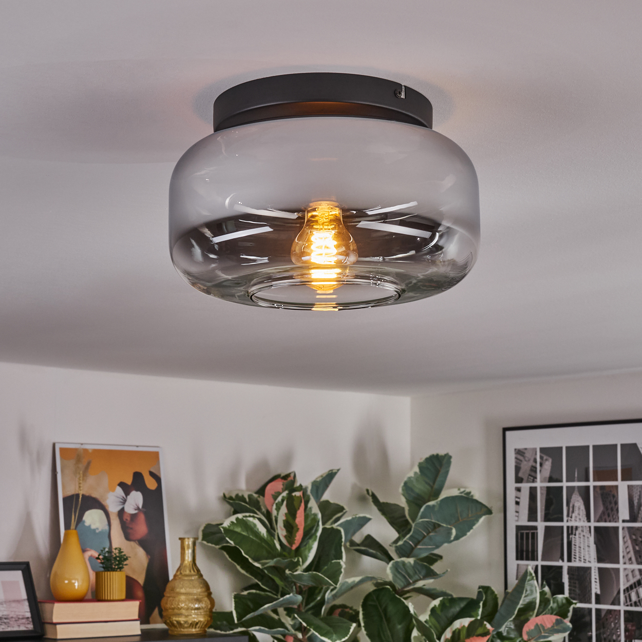 Eine moderne Deckenleuchte mit klarem Glasschirm, die einen Raum mit Kunst und Pflanzen beleuchtet.