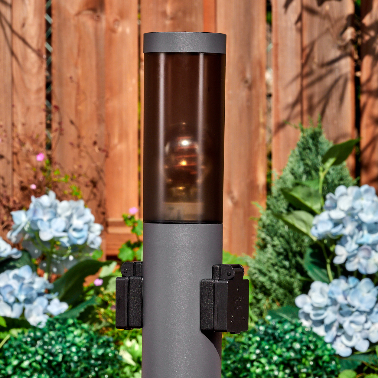 Graue Außenlampe mit braun getönter Abdeckung. Blaue Blumen und Grün umgeben die Lampe. Im Hintergrund ein Holzzaun.