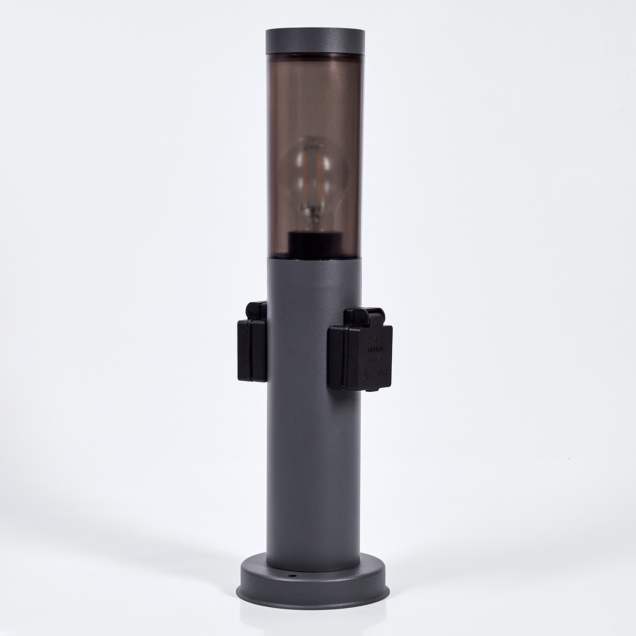 Eine graue Lampe auf weißer Oberfläche. Sie hat einen zylindrischen Körper, eine dunkle Oberseite mit einer Glühbirne und schwarze Seitenteile.