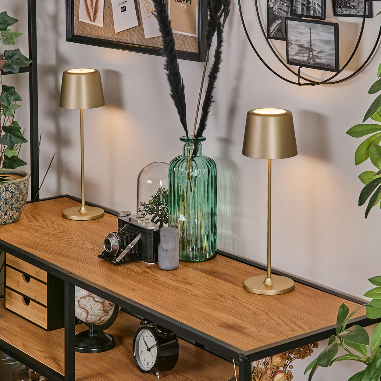 Zwei goldene Lampen auf einem Holzregal mit Dekoration wie Kamera, Uhr und grüner Vase. Hintergrund mit Bildern an der Wand.