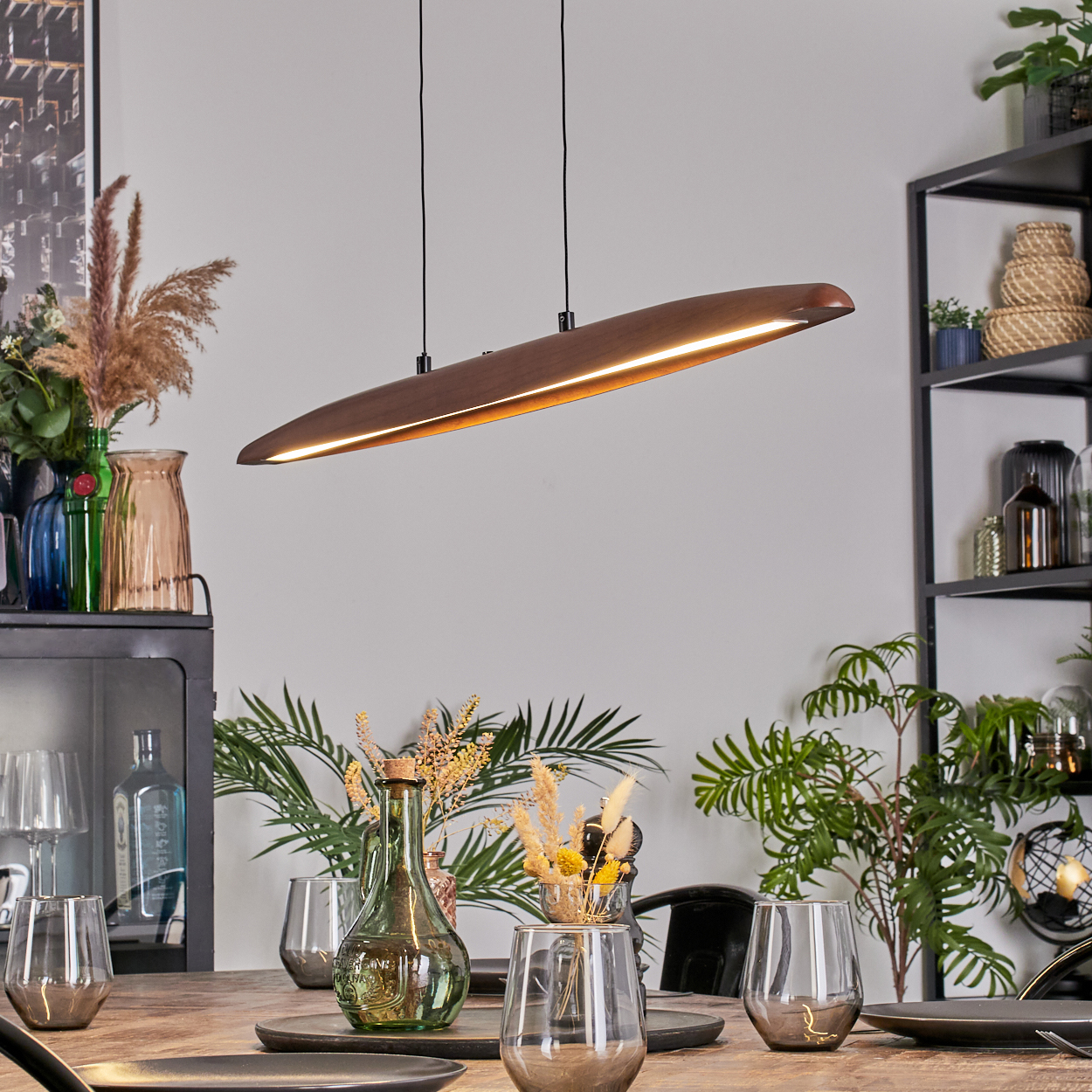 Ein Esstisch mit Gläsern, Tellern und einer Holzlampe. Pflanzen und Dekorationen füllen den Hintergrund.