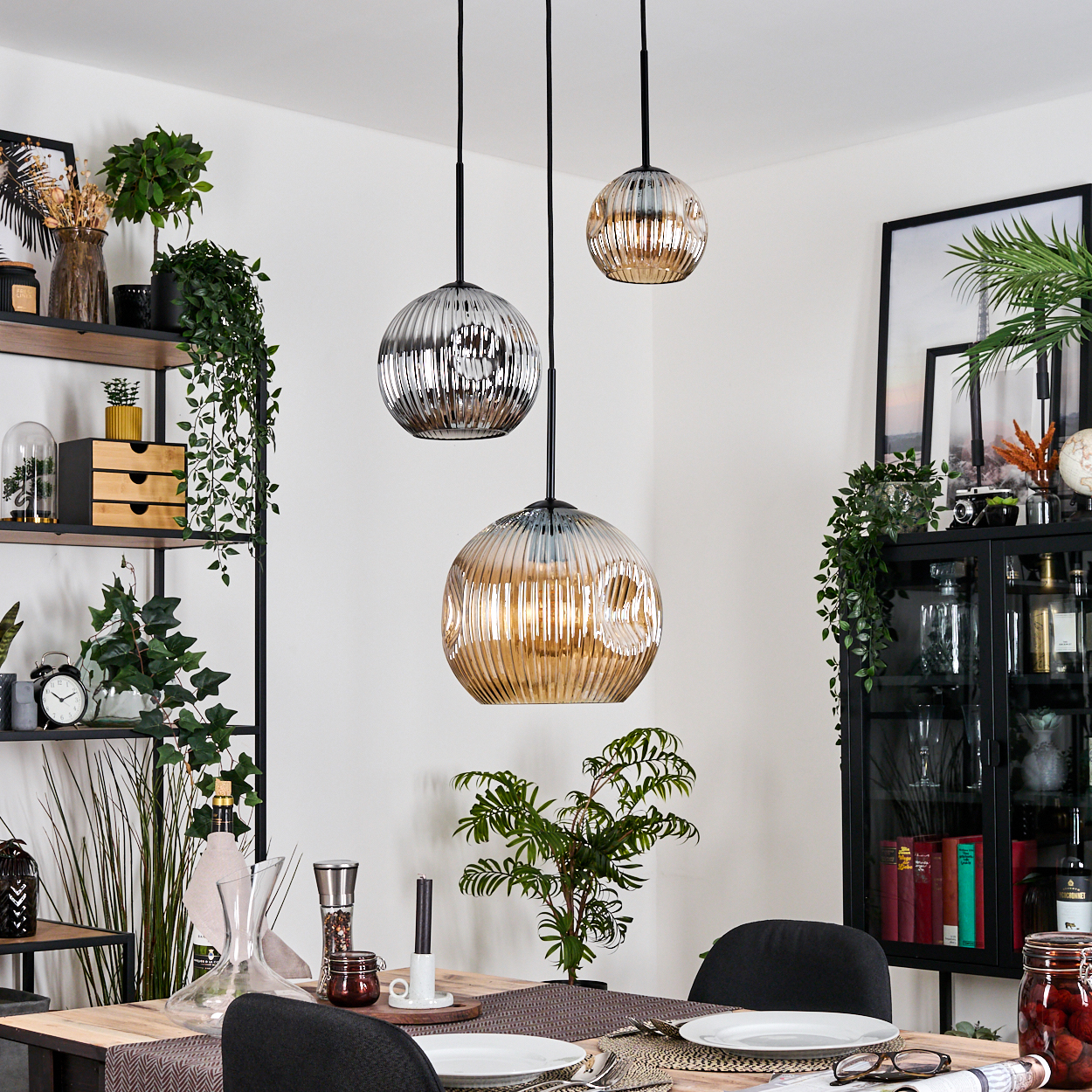 Esszimmer mit drei Hängelampen, gedecktem Tisch und dekorativen Elementen wie Pflanzen und einem Schrank.