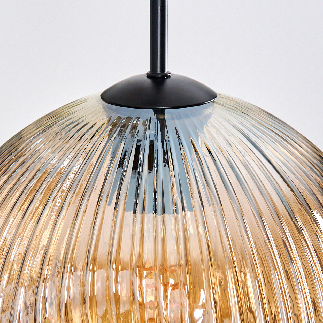 Nahaufnahme einer Pendelleuchte mit geriffeltem, bernsteinfarbenem Glasschirm. Die Leuchte hat einen schwarzen Metallschaft und einen weißen Hintergrund.