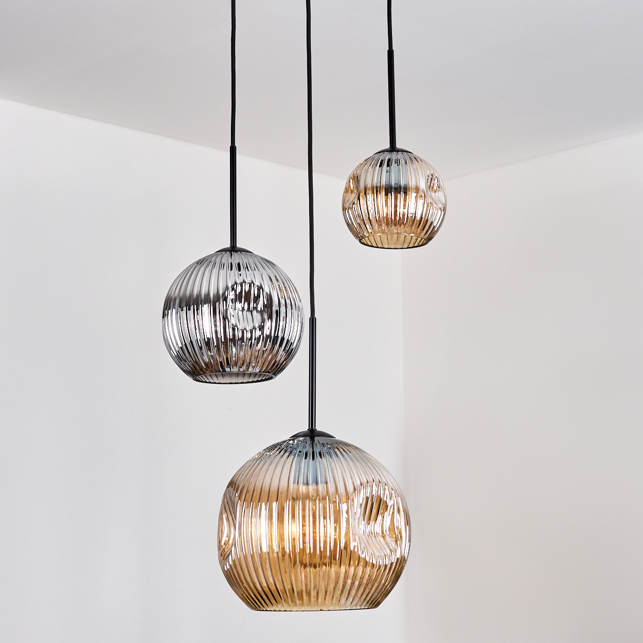 Drei kugelförmige Pendelleuchten mit geriffeltem Glas hängen an schwarzen Schnüren vor einer weißen Wand und Decke. Die Lichtfarben variieren.