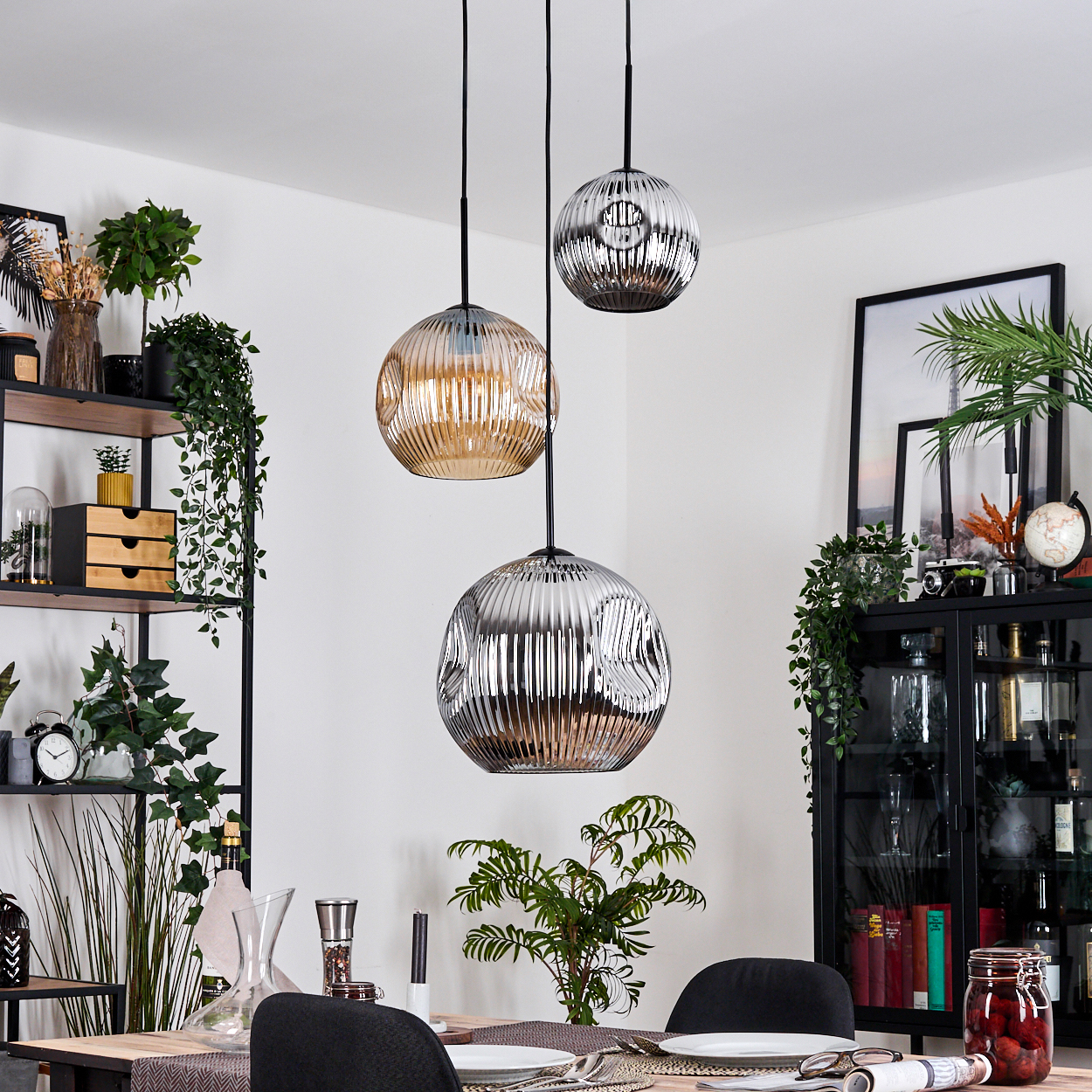 Esszimmer mit drei Hängelampen. Tisch zum Essen gedeckt. Dekorative Pflanzen und Regale mit verschiedenen Gegenständen.