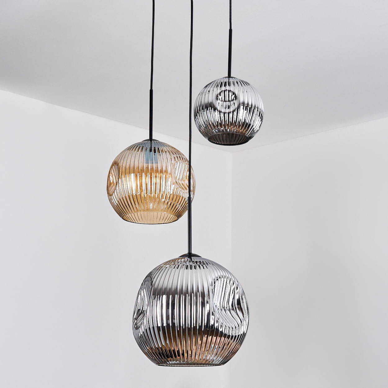 Drei kugelförmige Pendelleuchten hängen von der Decke. Zwei sind metallisch, eine ist bernsteinfarben, alle mit strukturierter Oberfläche. Der Hintergrund ist weiß.
