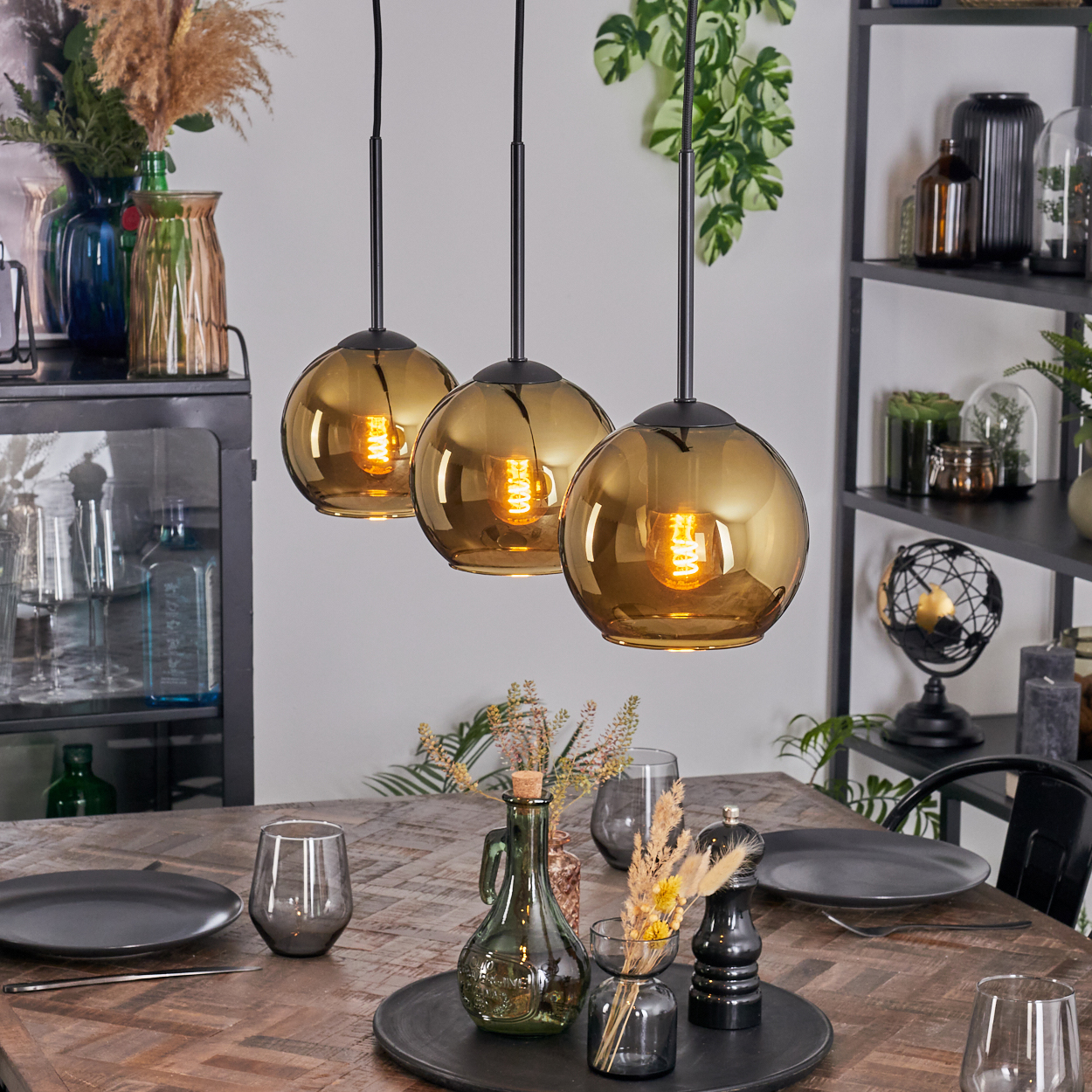 Drei goldene Pendelleuchten über einem Holztisch, der für das Abendessen gedeckt ist. Auf dem Tisch und in Regalen im Hintergrund stehen Dekorationsartikel.