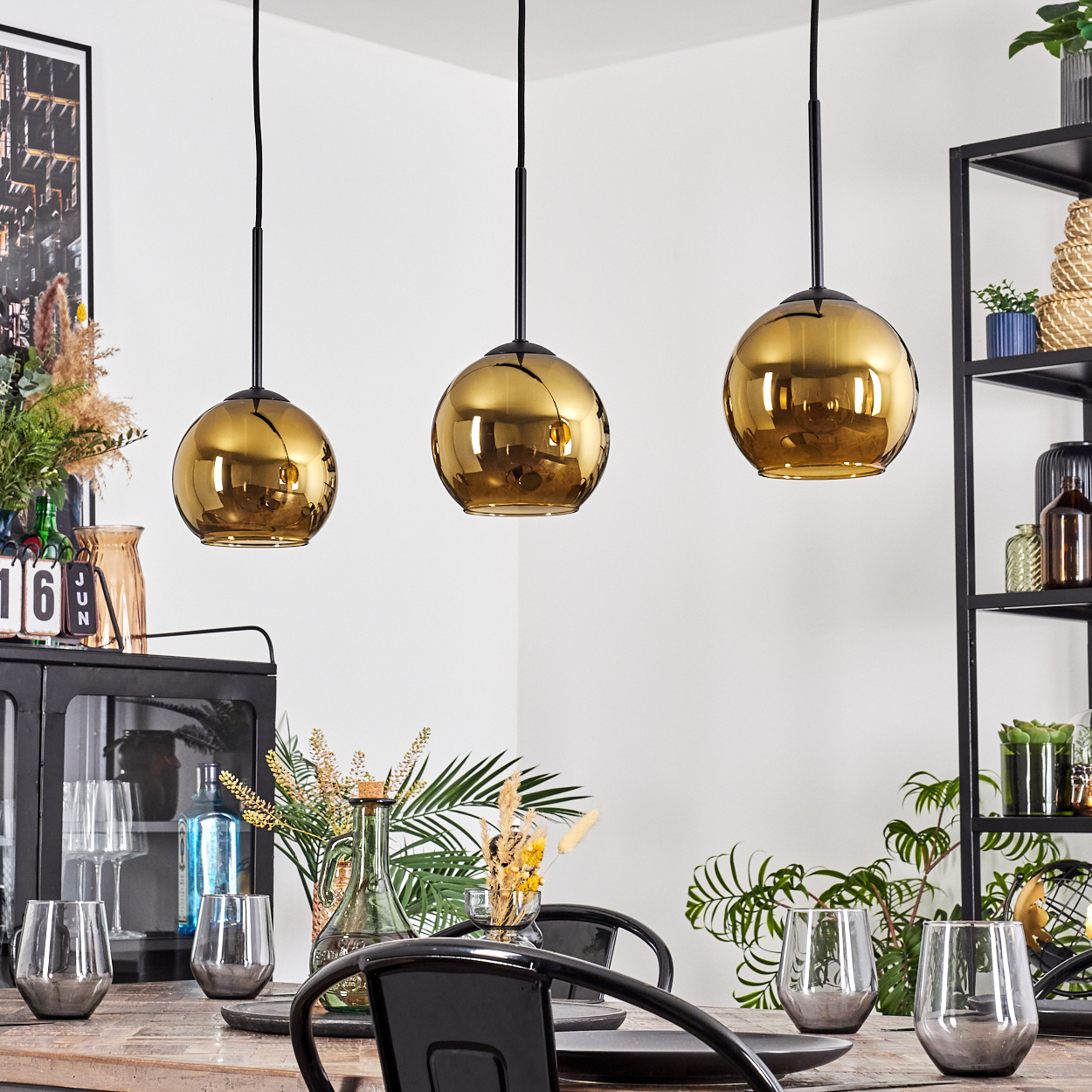 Drei goldene Pendelleuchten über einem Esstisch mit Gläsern und Tellern. Schwarze Metallstühle und ein Regal im Hintergrund.