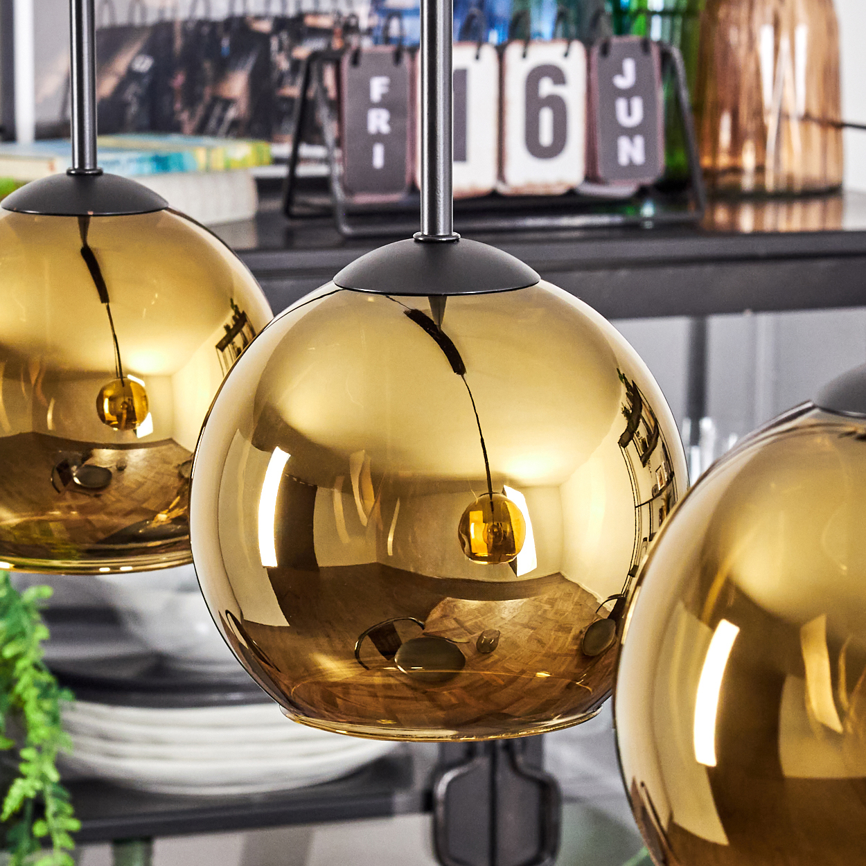 Drei goldene, kugelförmige Pendelleuchten. Die Leuchten reflektieren den Raum. Im Hintergrund befinden sich ein Kalender und Glaswaren.