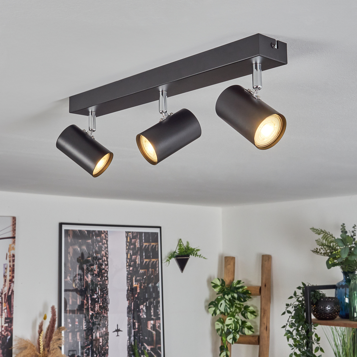 Drei schwarze Strahler an der Decke befestigt. Die Lichter sind eingeschaltet und leuchten nach unten. Im Hintergrund ein Bild und Pflanzen.